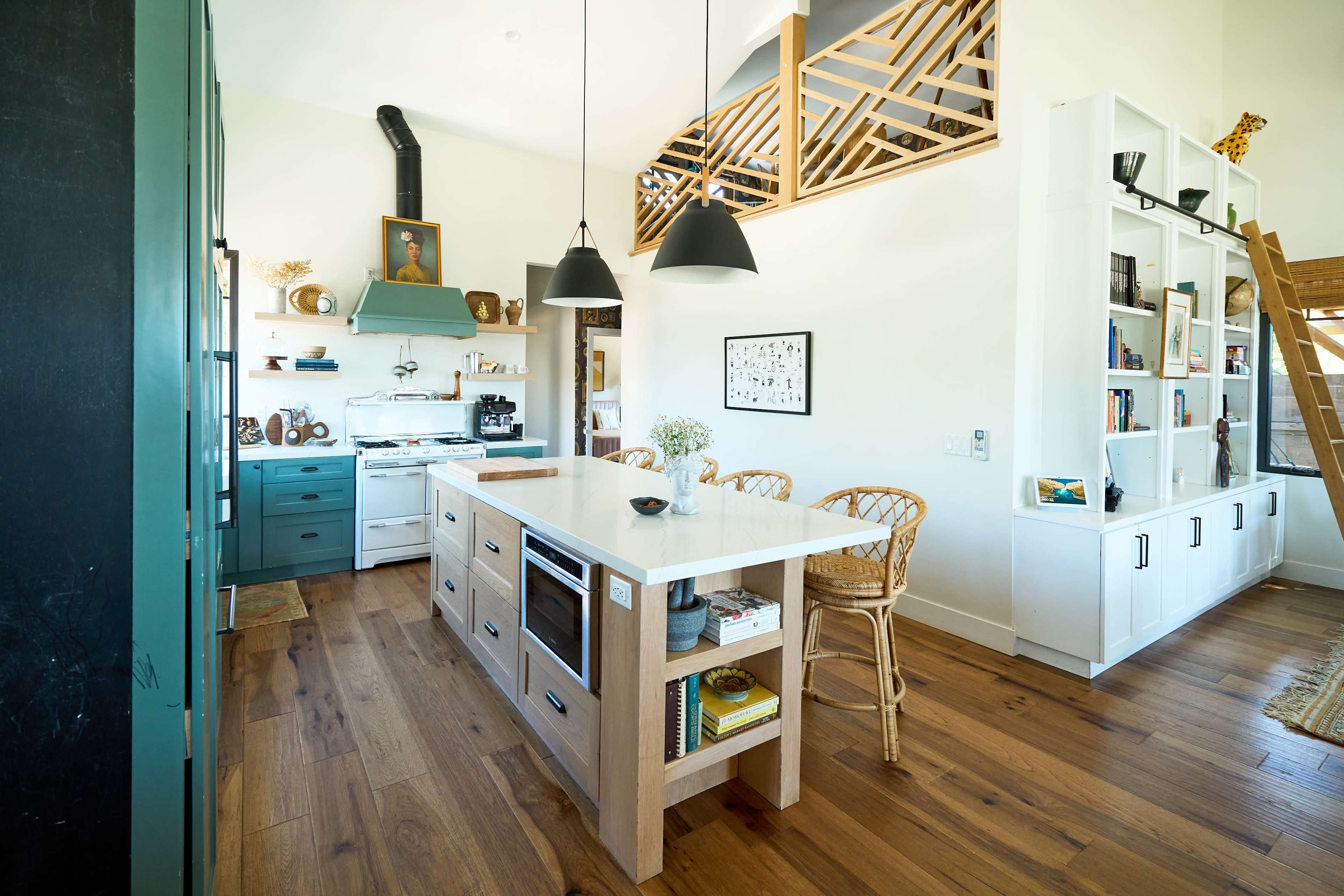 The image shows a modern kitchen with a central island, featuring light wood cabinetry, a blue stove, and open shelving, adjacent to a living area with bookshelves and decorative items.