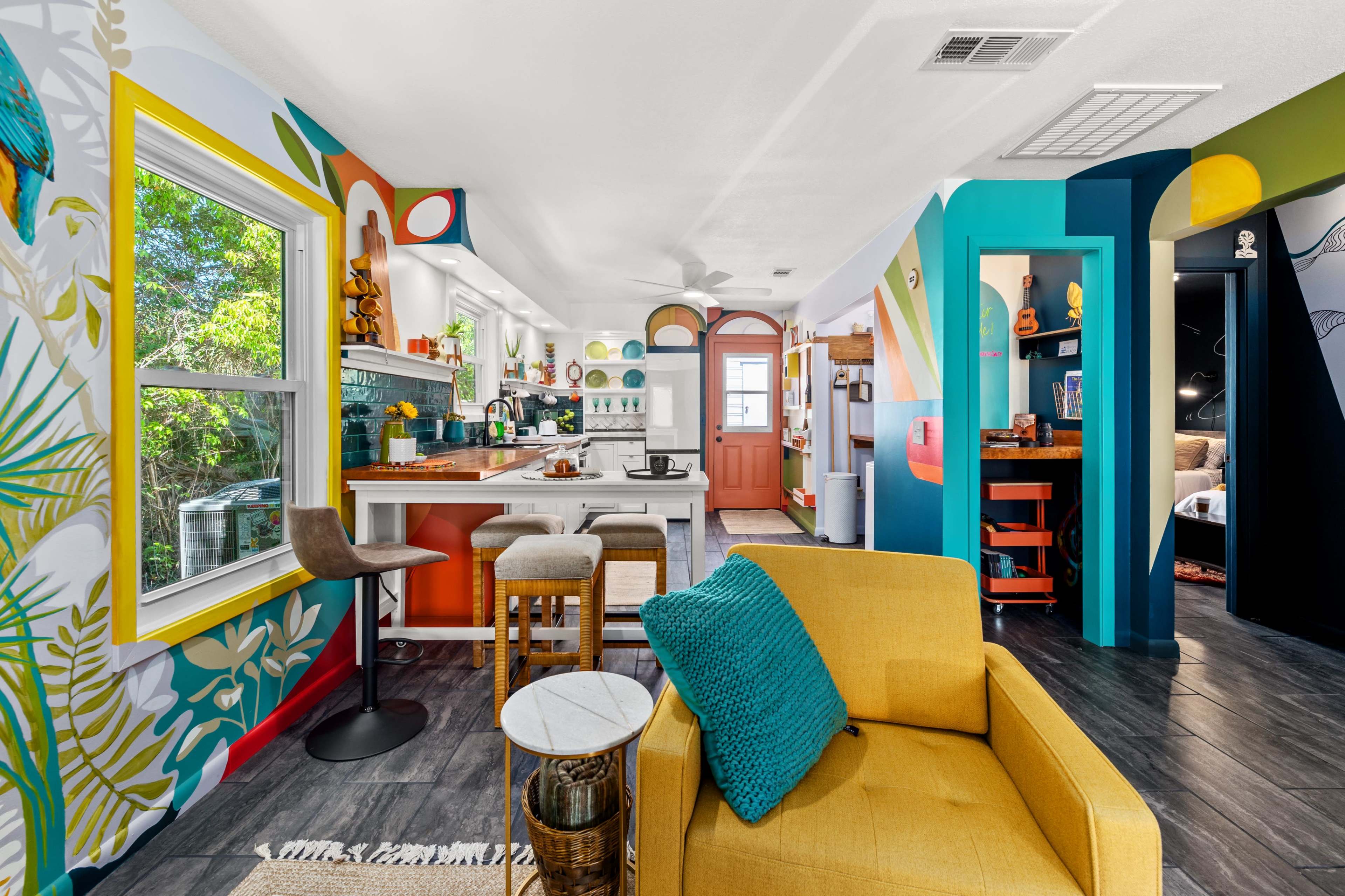 A brightly colored kitchen and living area featuring eclectic decor, a yellow armchair, and vibrant wall murals.