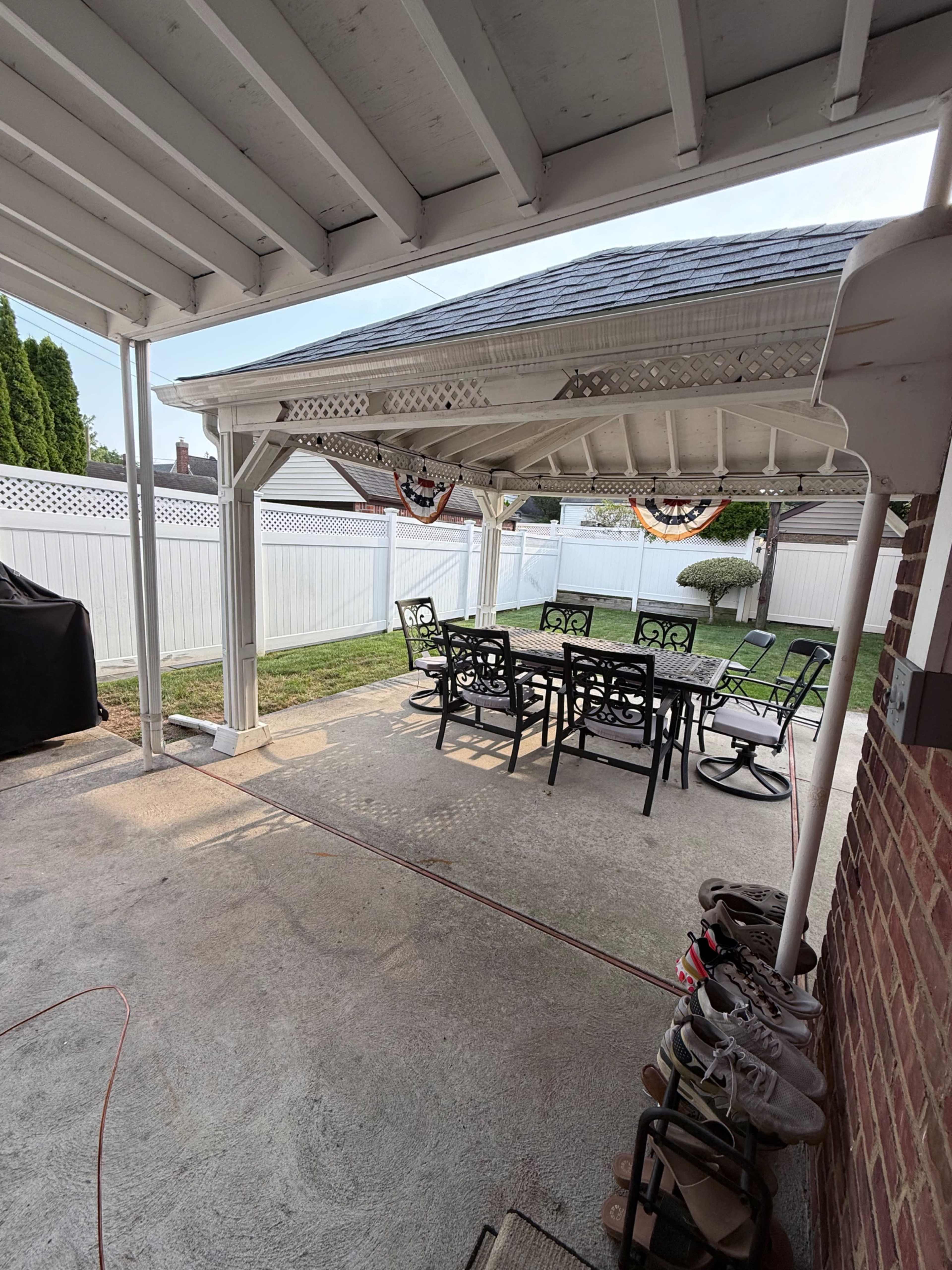 The image shows a covered patio area with a table and chairs, overlooking a lawn with a white fence in the background.