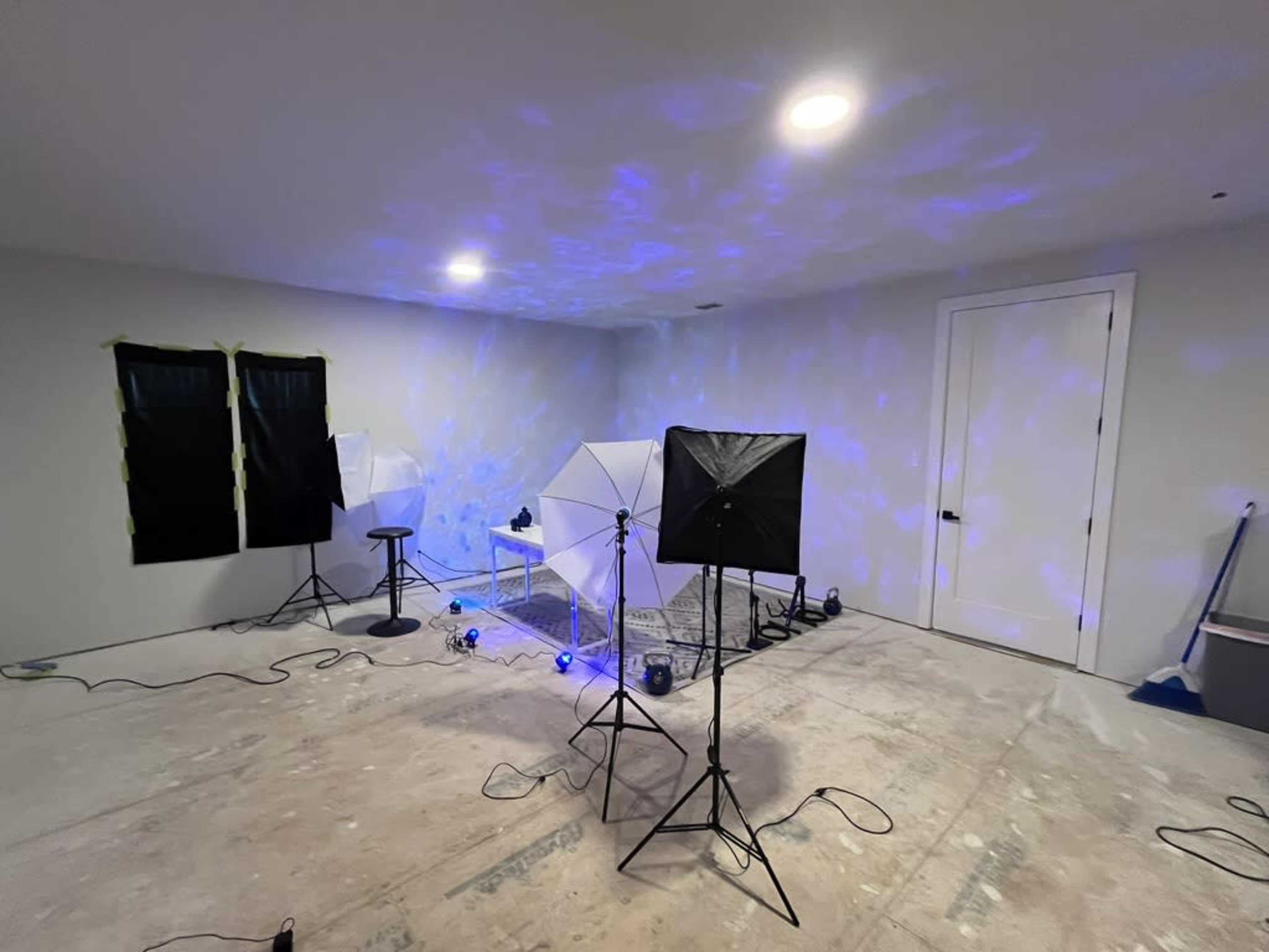 The image shows a sparsely furnished room with lighting equipment set up, including two softbox lights and a stool, on an unfinished floor.