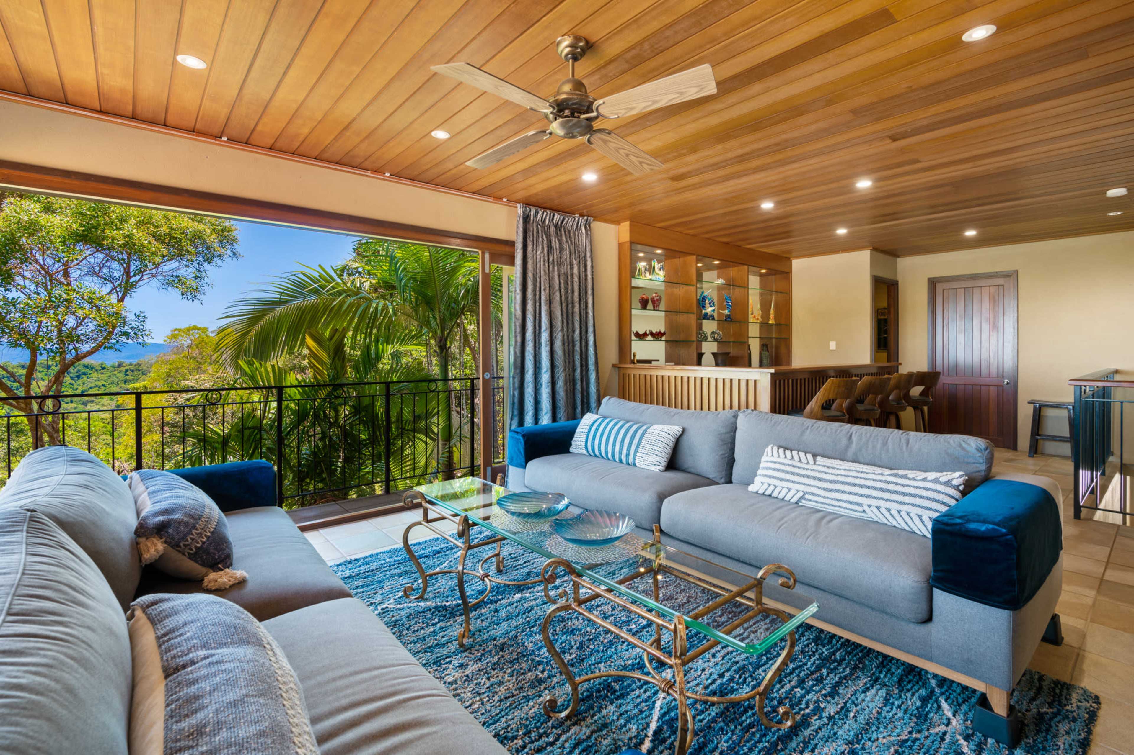 A modern living room features large windows with a view of nature, a comfortable seating area with blue and gray sofas, and a decorative coffee table under wooden ceilings.