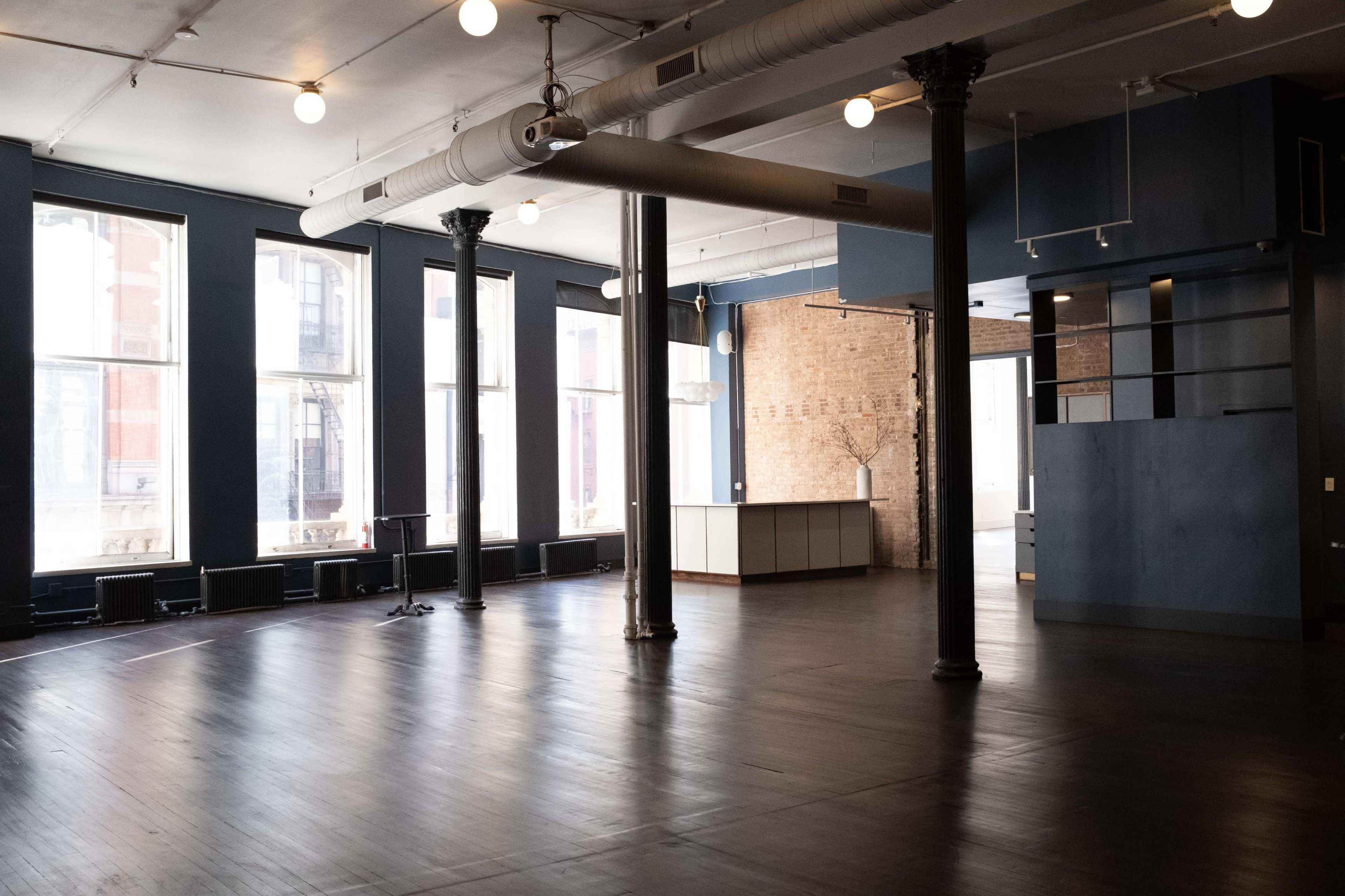 The image shows a spacious, empty room with large windows, wooden floors, exposed brick walls, and columns.