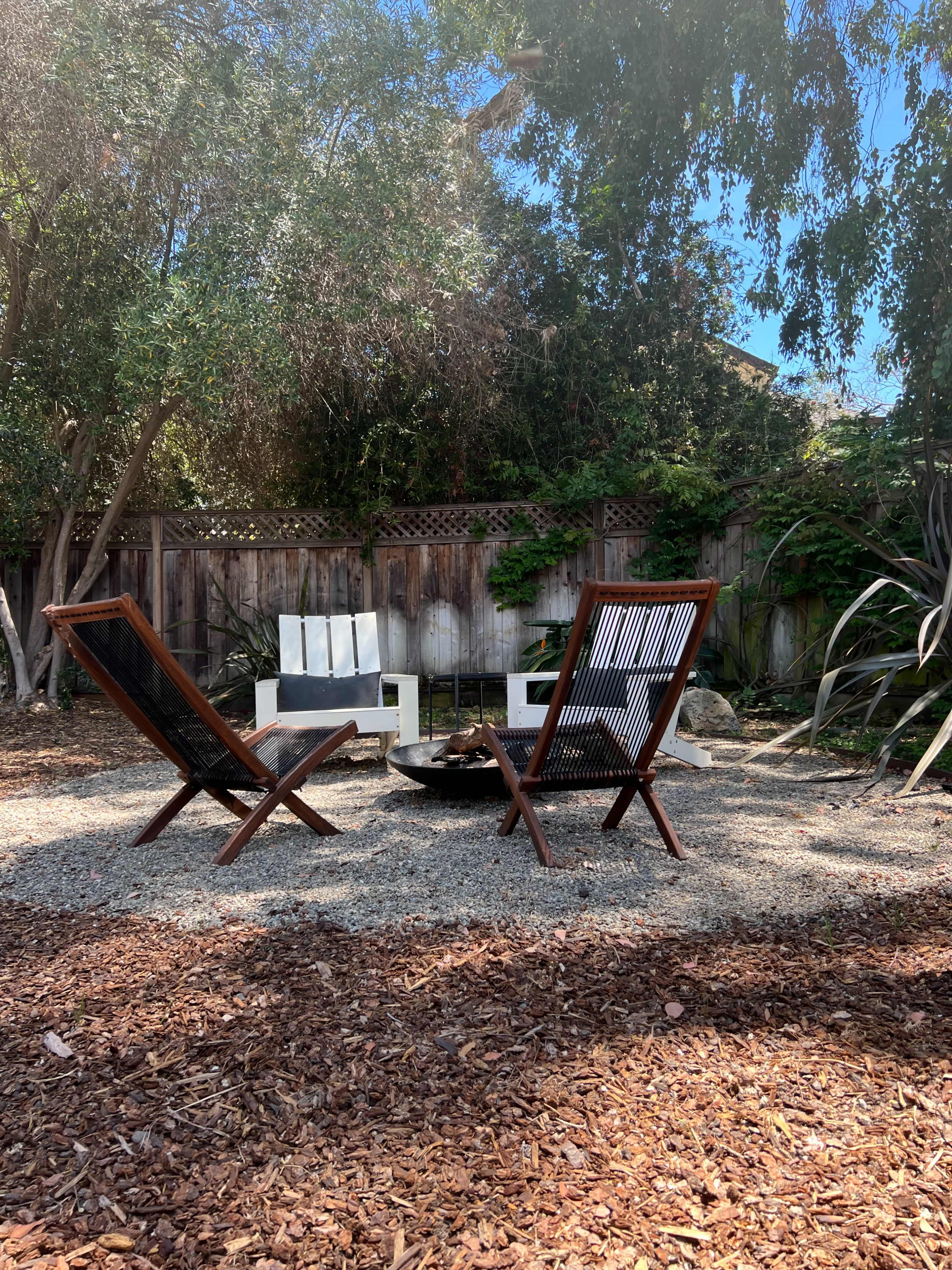 Two wooden chairs face a circular fire pit in a gravelly backyard surrounded by trees and a wooden fence.