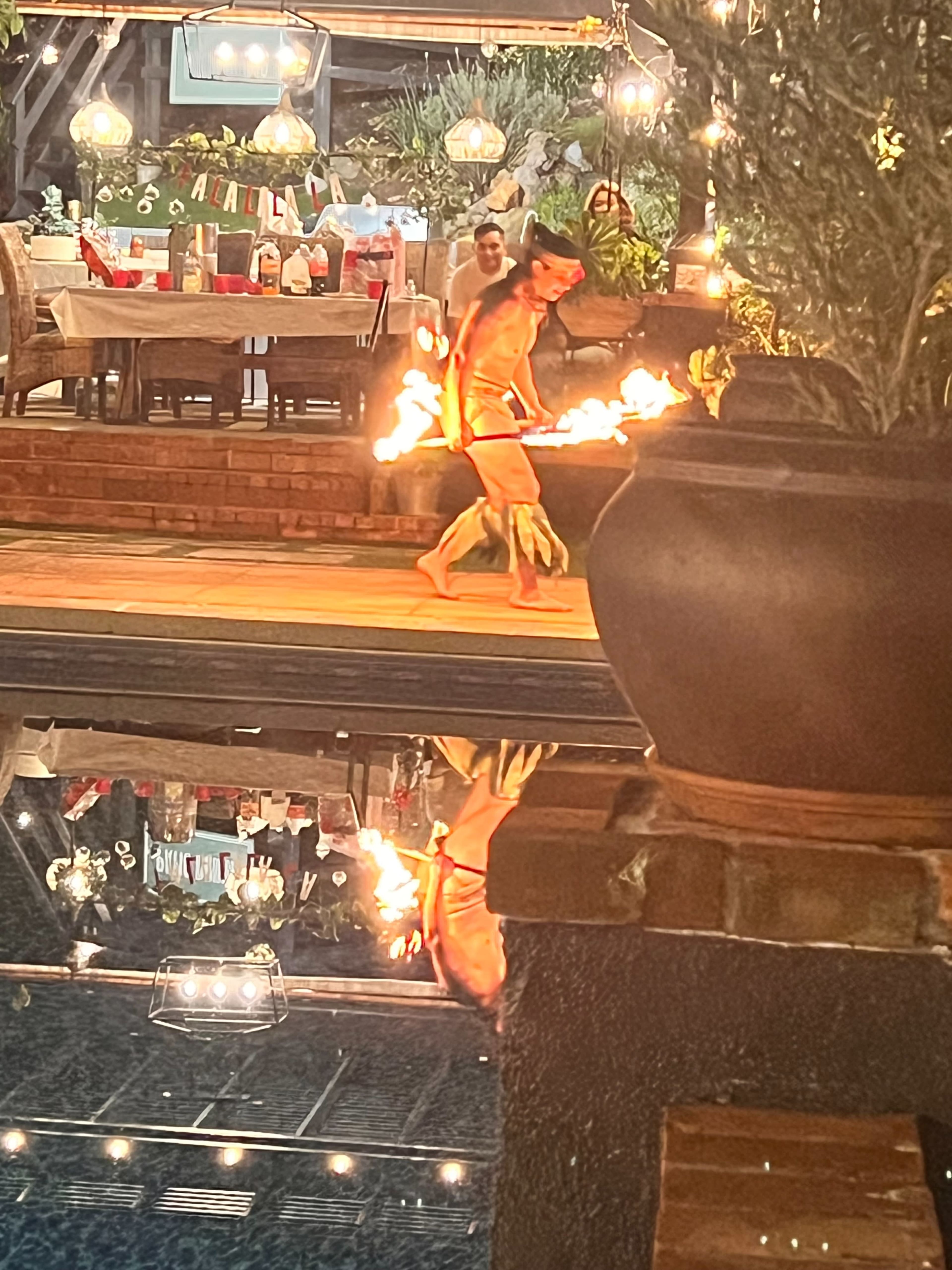 A performer twirls flaming torches while walking by a swimming pool at a well-decorated outdoor venue.