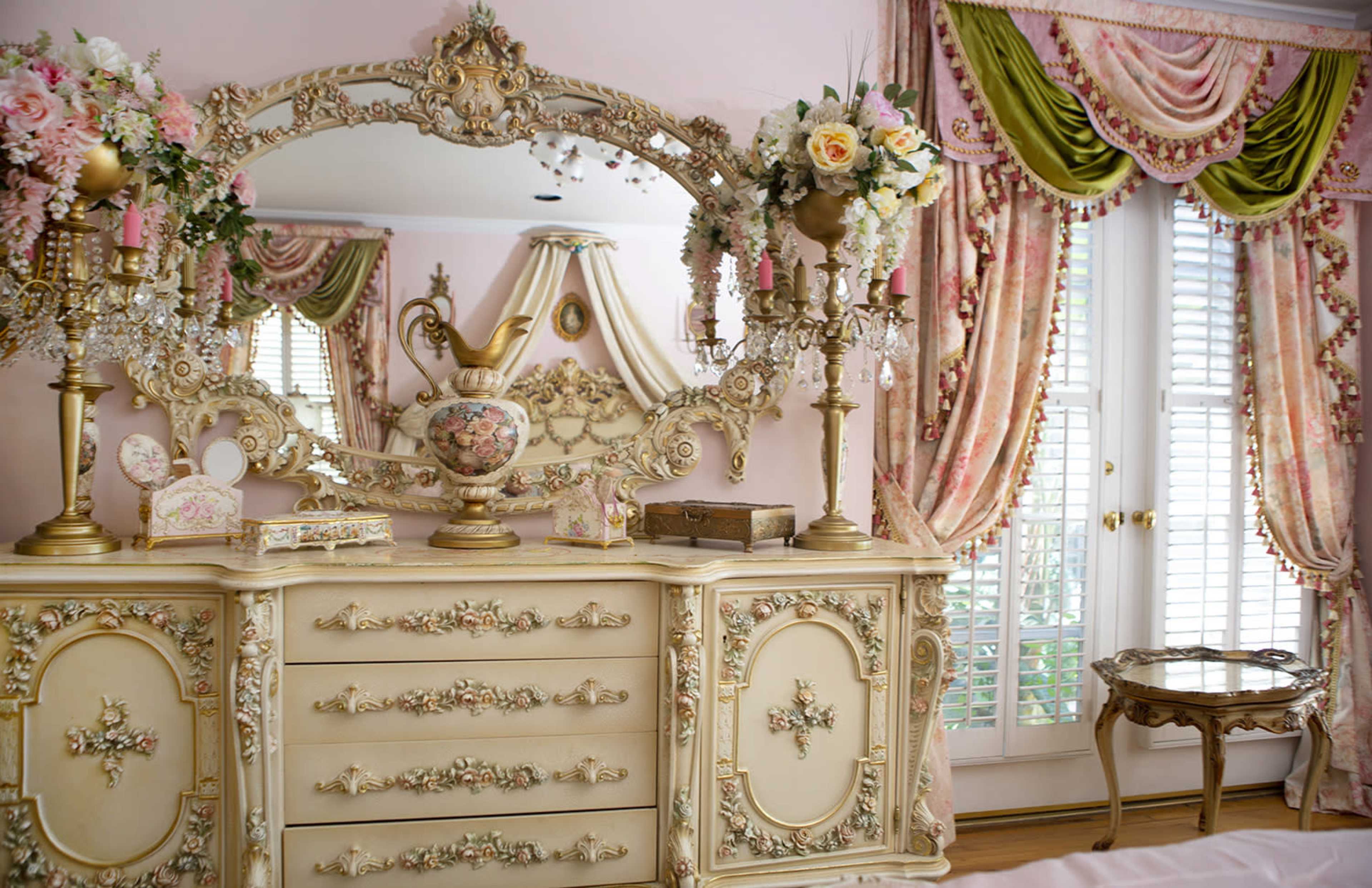A decorated bedroom featuring an ornate dresser with floral designs, a large mirror, and draped curtains.