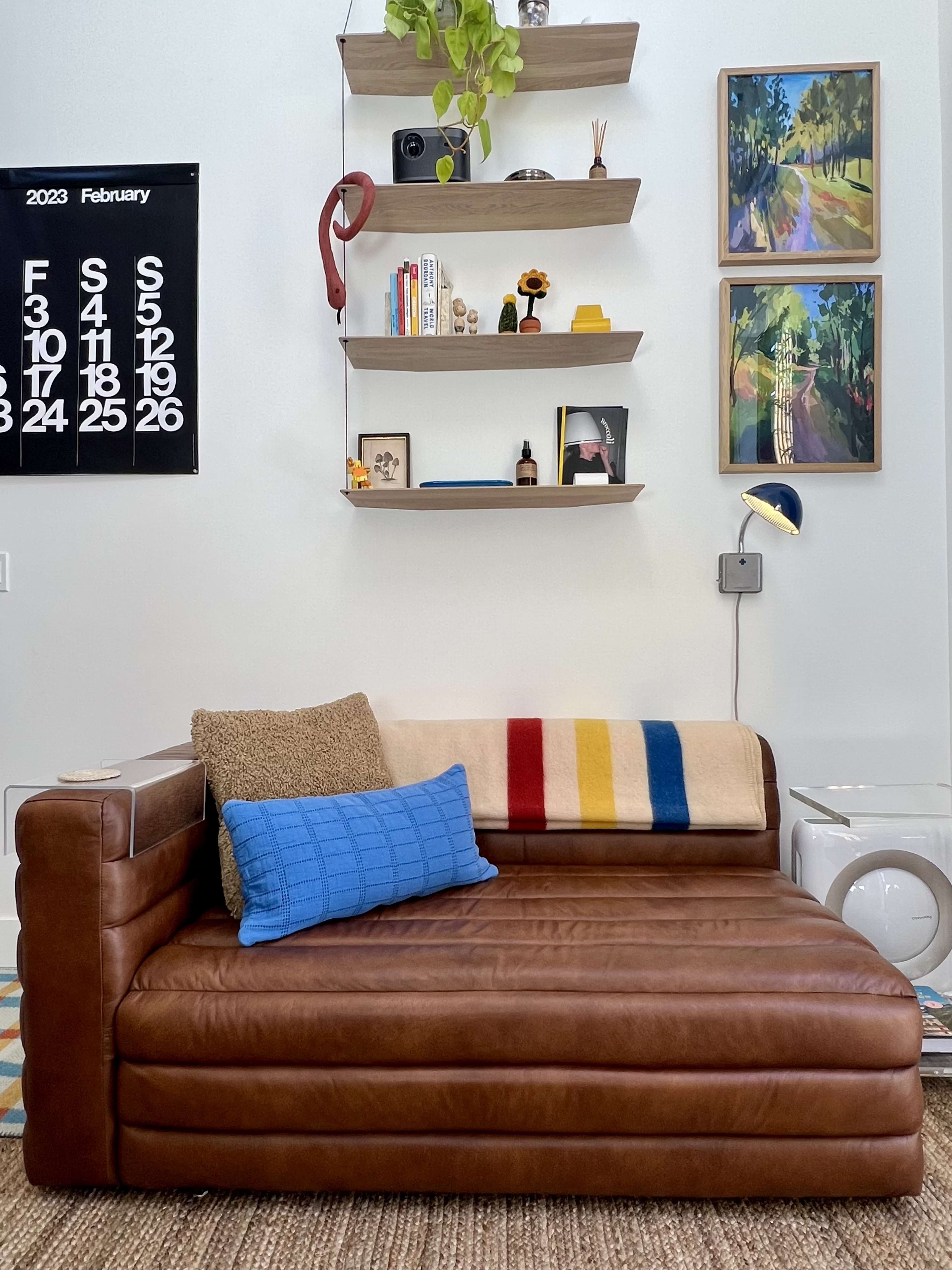 A brown leather sofa with a blue pillow and a striped throw sits against a wall adorned with shelves and framed artwork.