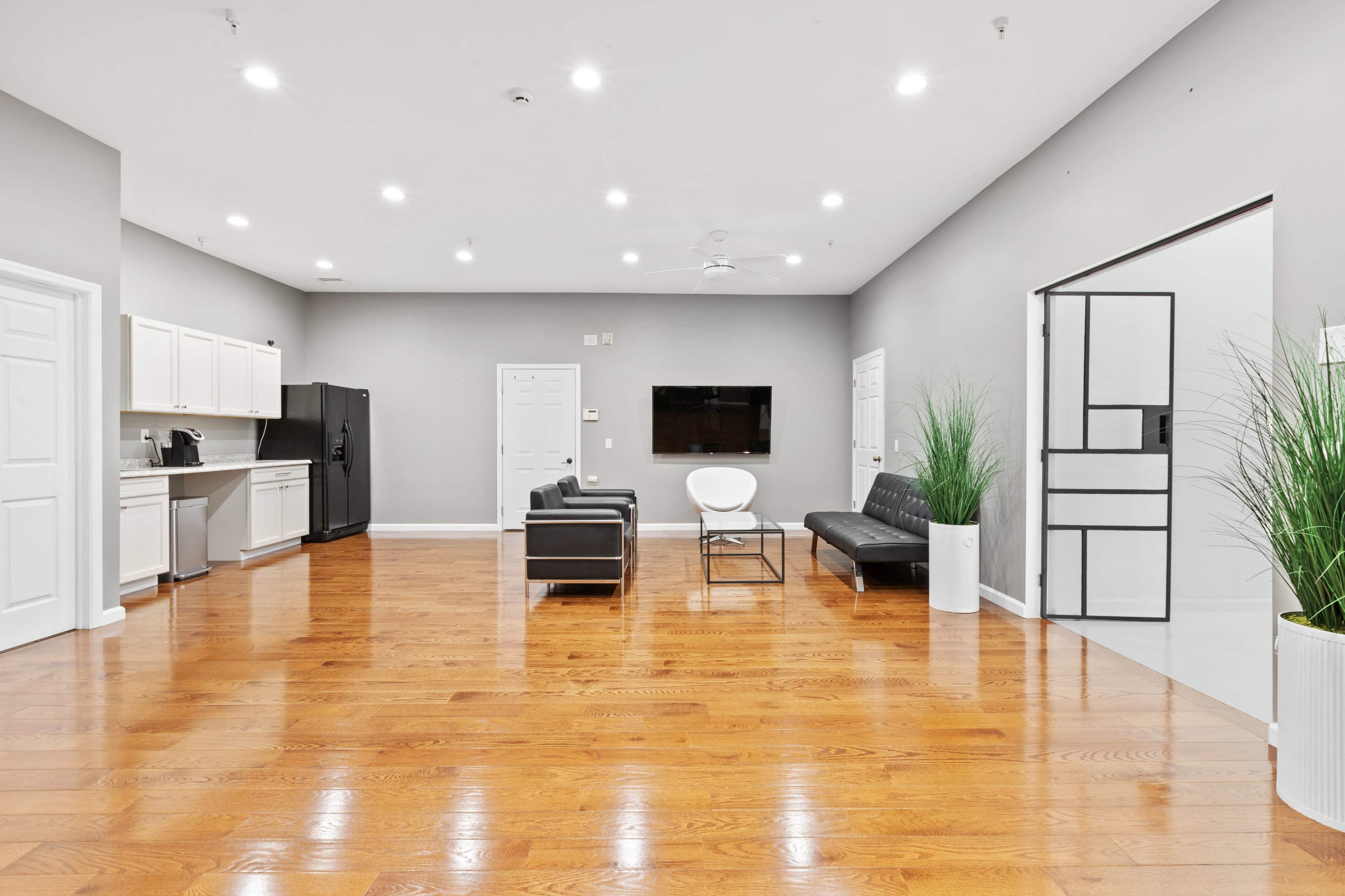 A spacious, modern room features hardwood flooring, a seating area with two black chairs, a white chair, and a wall-mounted television, alongside kitchen cabinets and appliances.