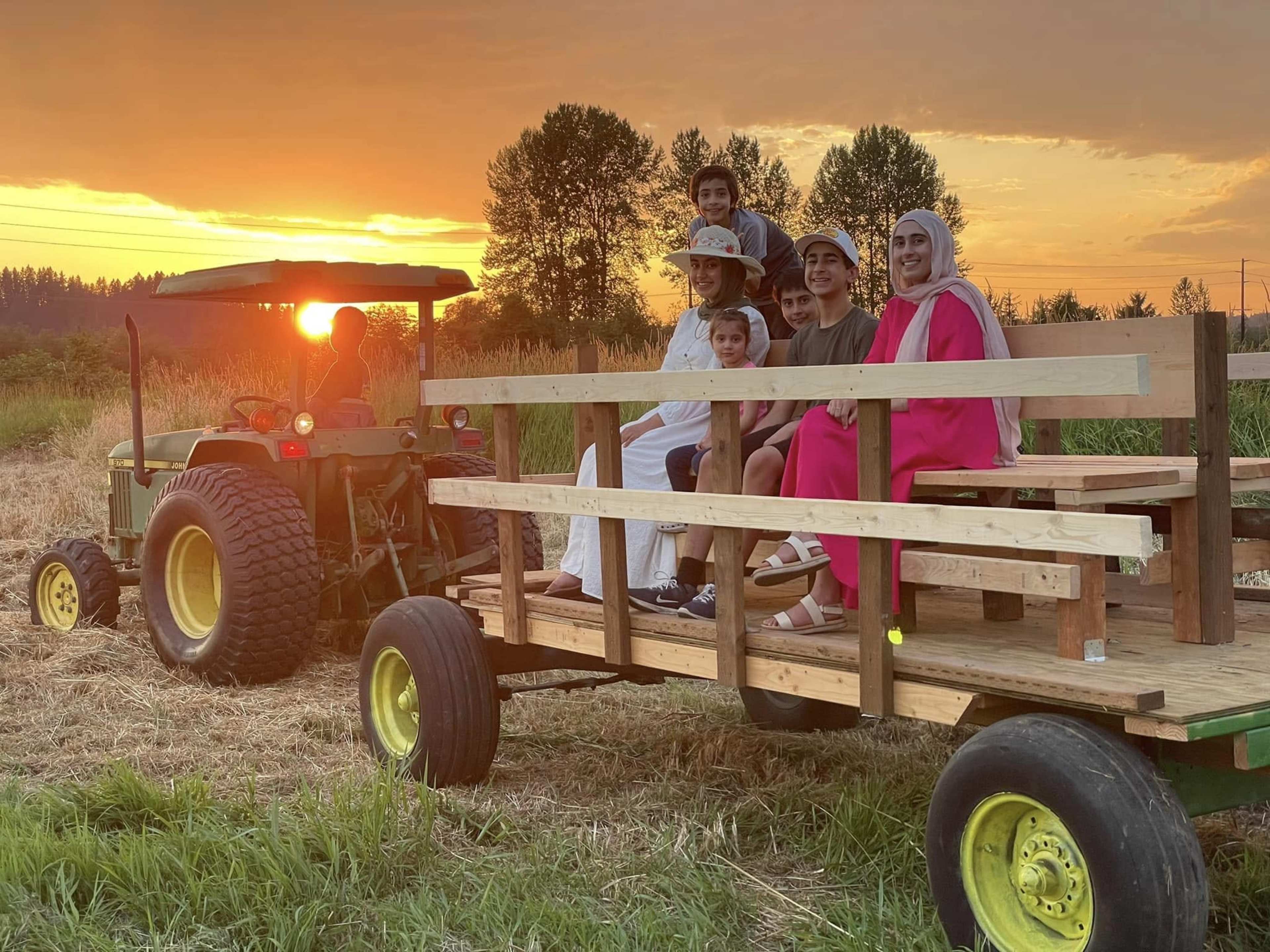A family sits on a wooden wagon attached to a tractor during a sunset in a field.