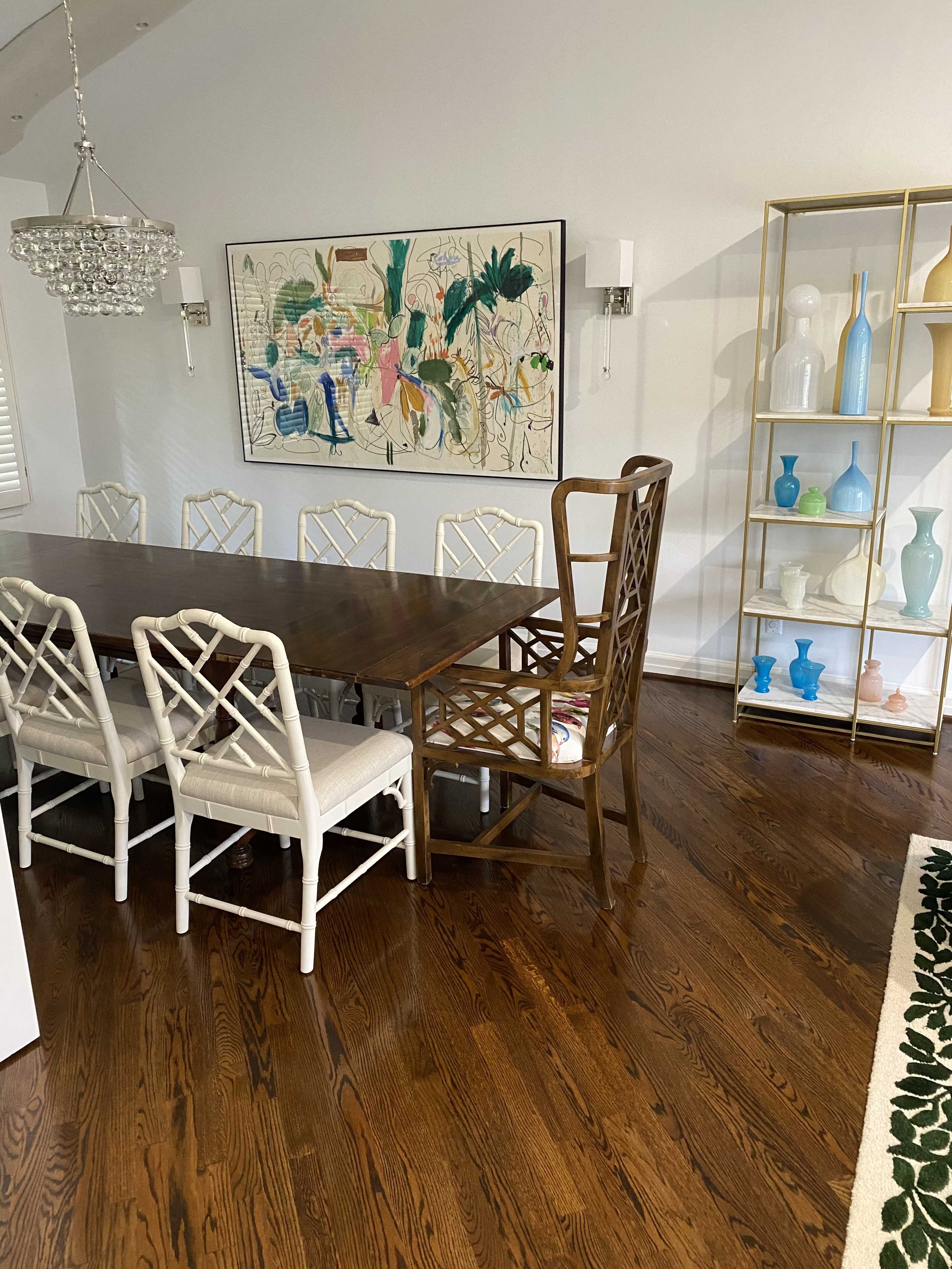 A dining room features a long wooden table surrounded by white chairs, with a large wall art piece and a glass display shelf filled with various decorative items.
