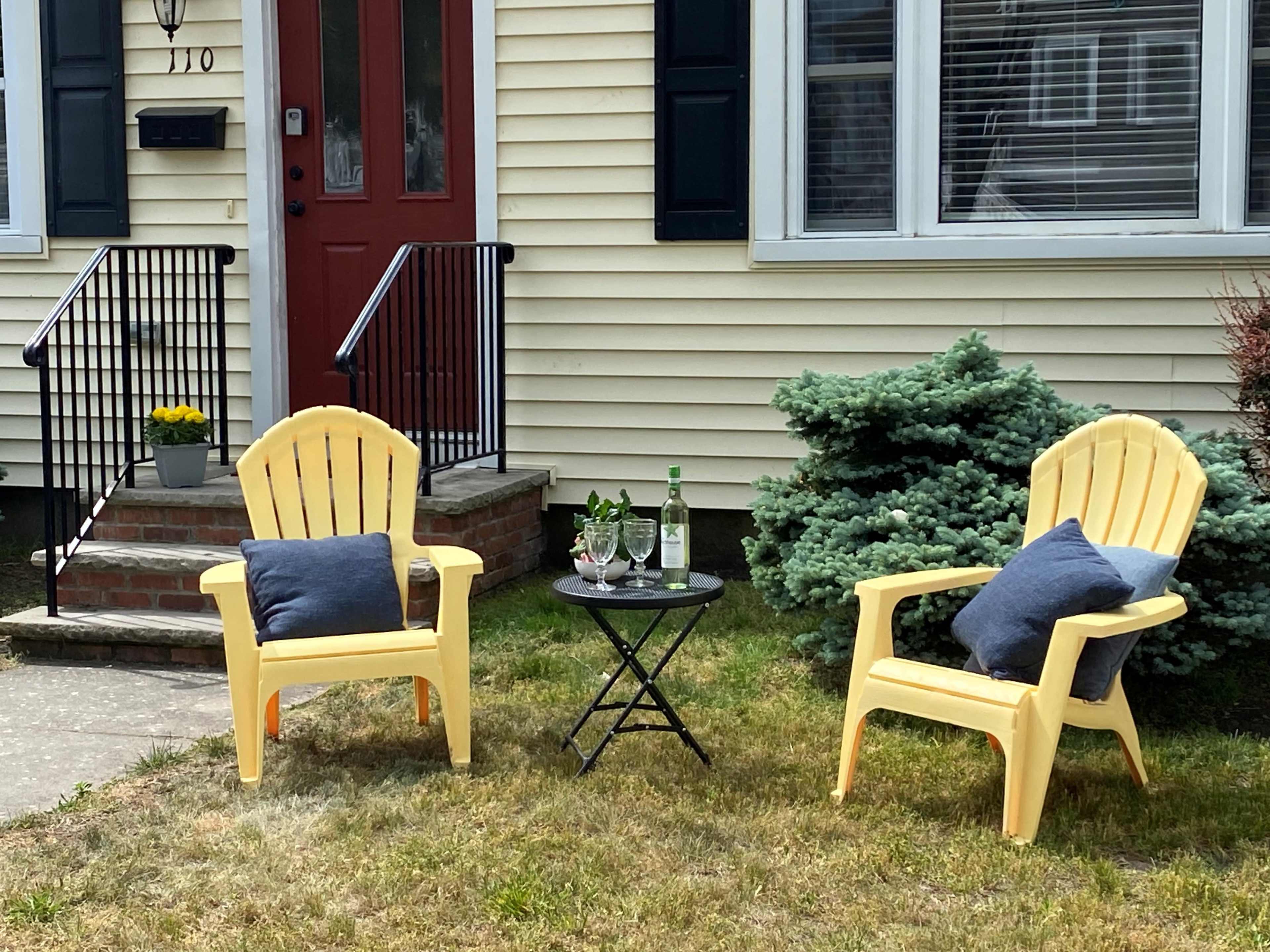 Two yellow Adirondack chairs with blue cushions flank a small table featuring glasses and a bottle, set on a grassy area in front of a house.