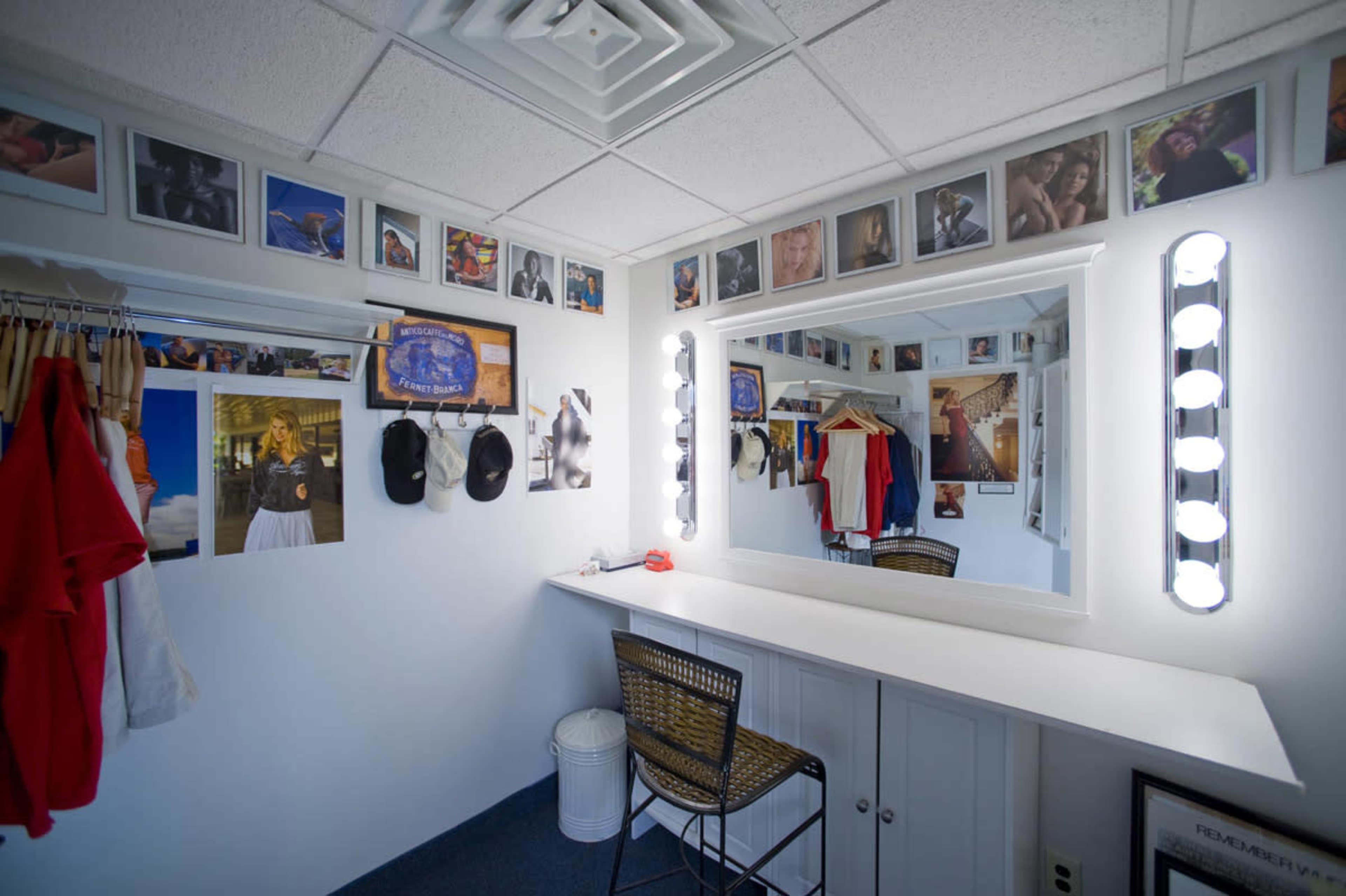 A dressing room with a large mirror, bright light fixtures, and various clothing items and photographs displayed on the walls.
