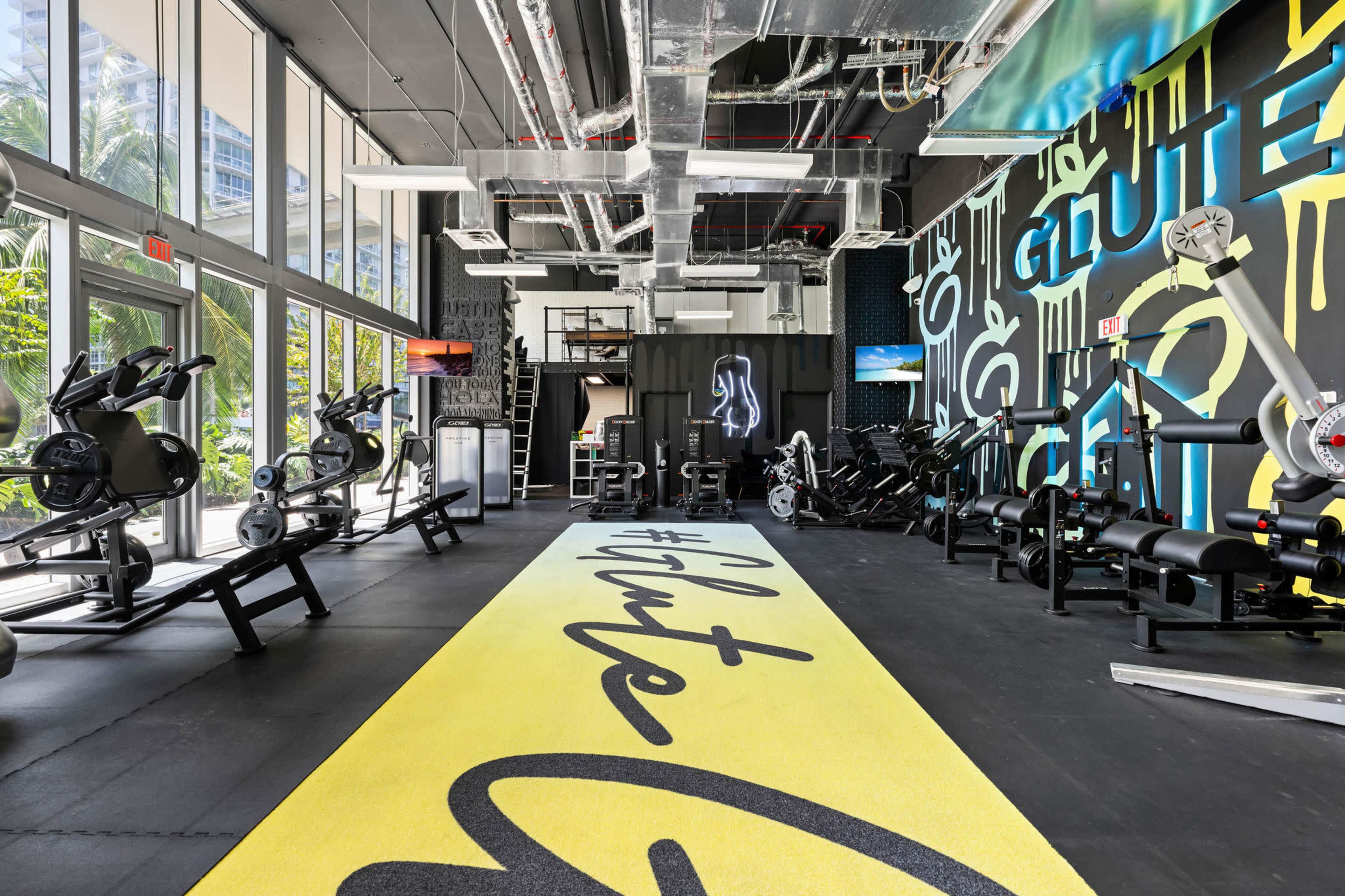 A bright gym interior featuring exercise equipment and a yellow carpet with the word "Glute" prominently displayed.
