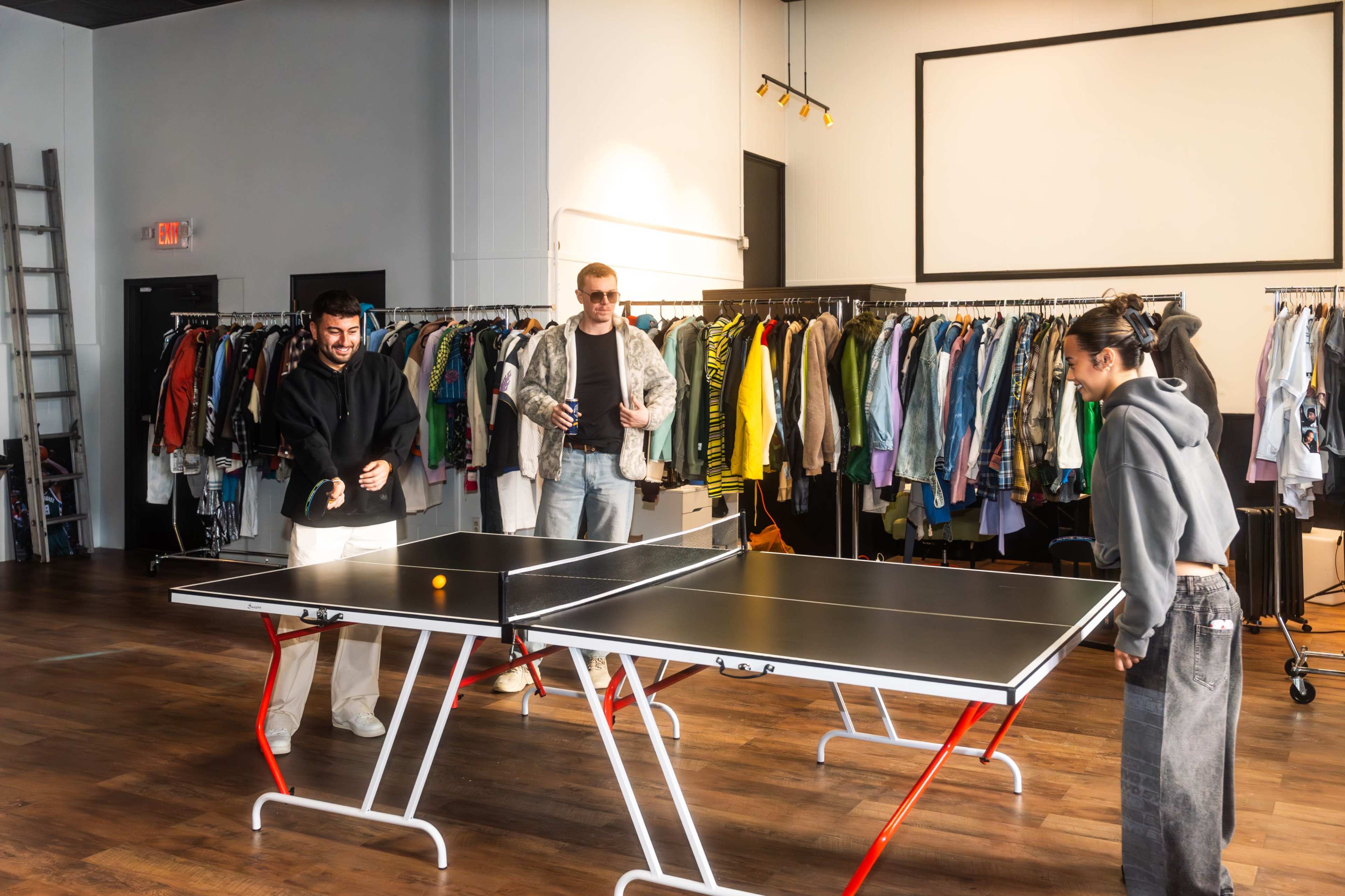 Three people play table tennis in a clothing store with racks of clothes in the background.
