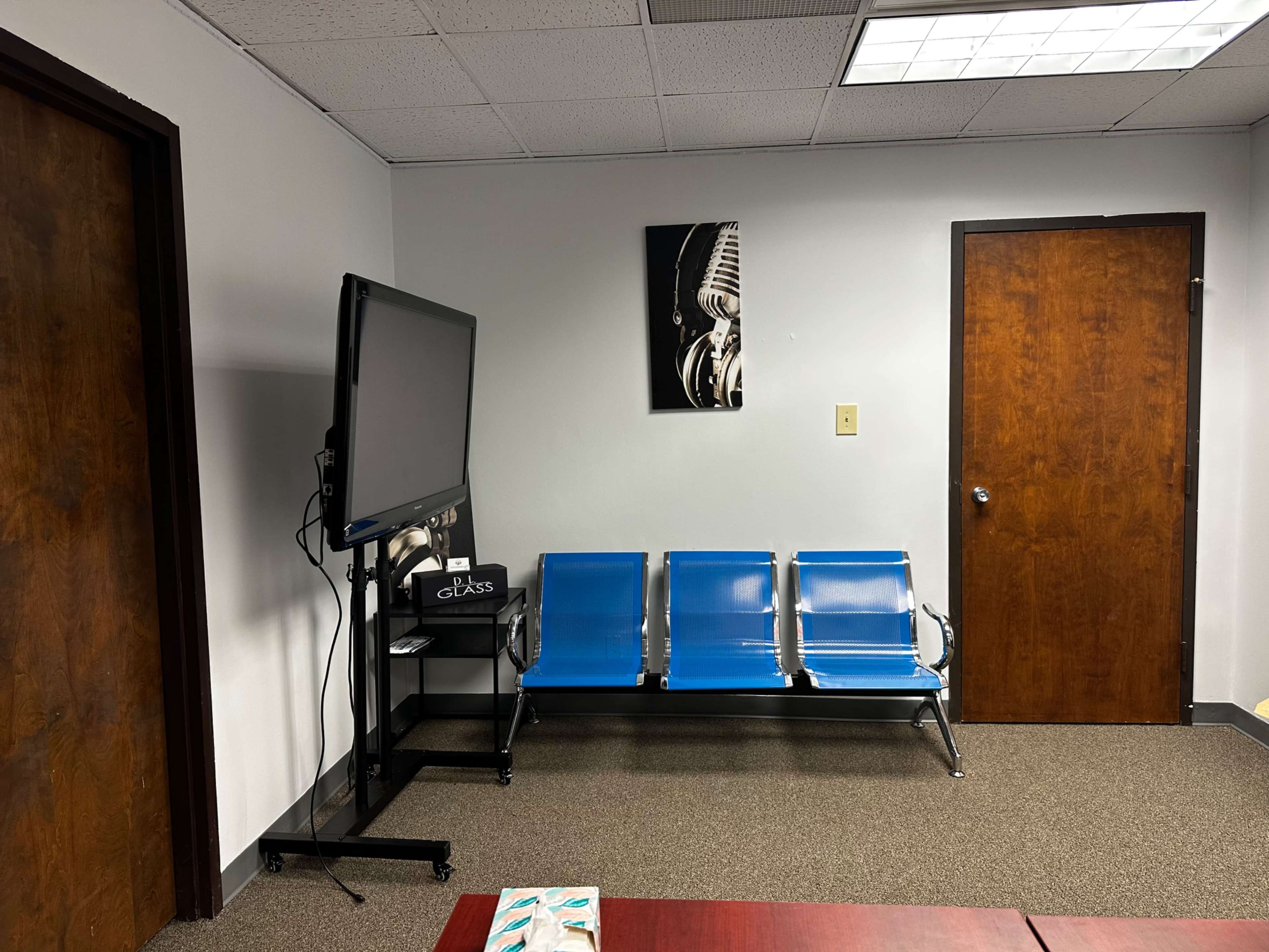 The image shows a waiting area with three blue chairs, a television on a stand, and a framed black and white artwork of a guitar on the wall.