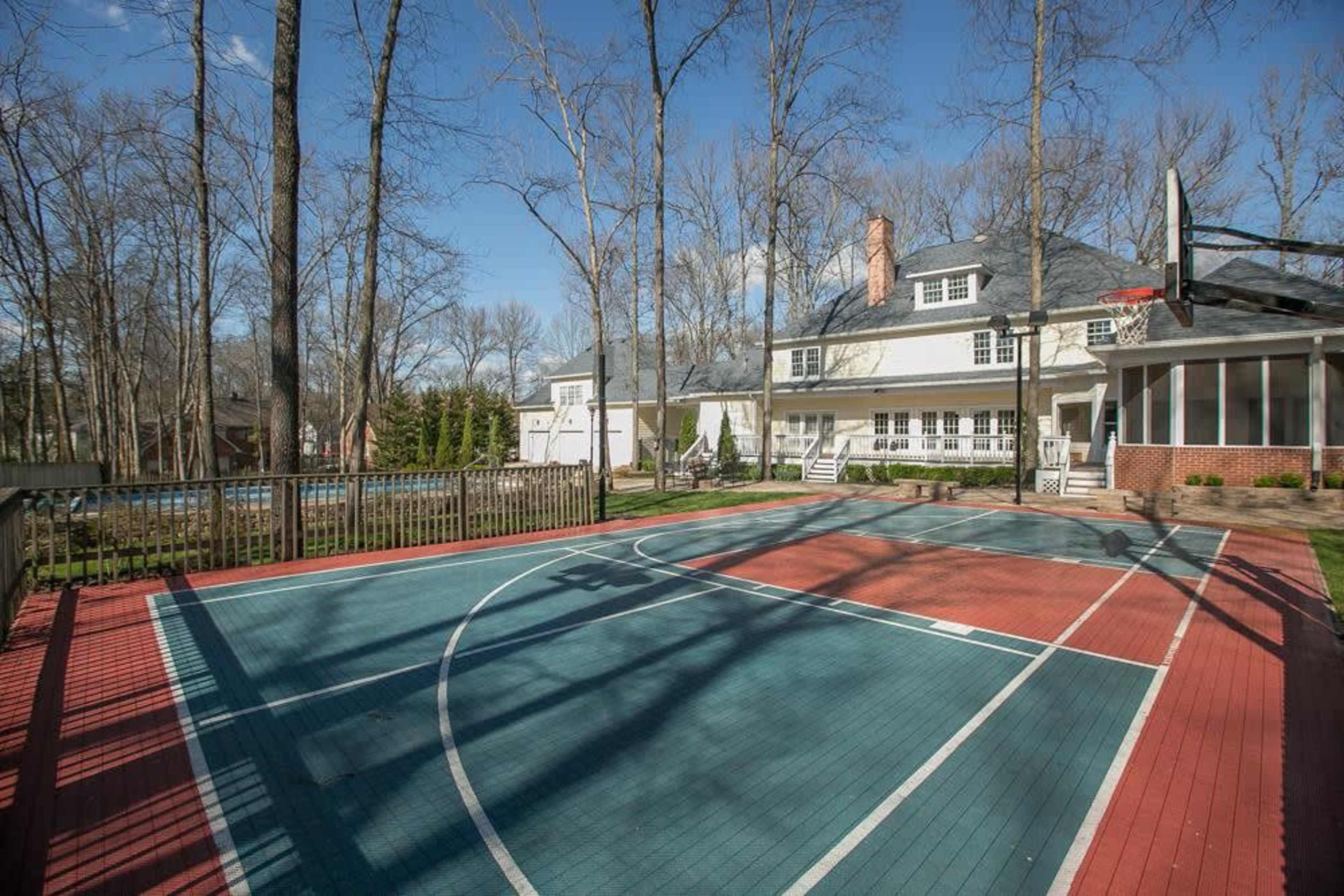 A basketball court with red and turquoise surfaces in a backyard surrounded by trees and a large house.