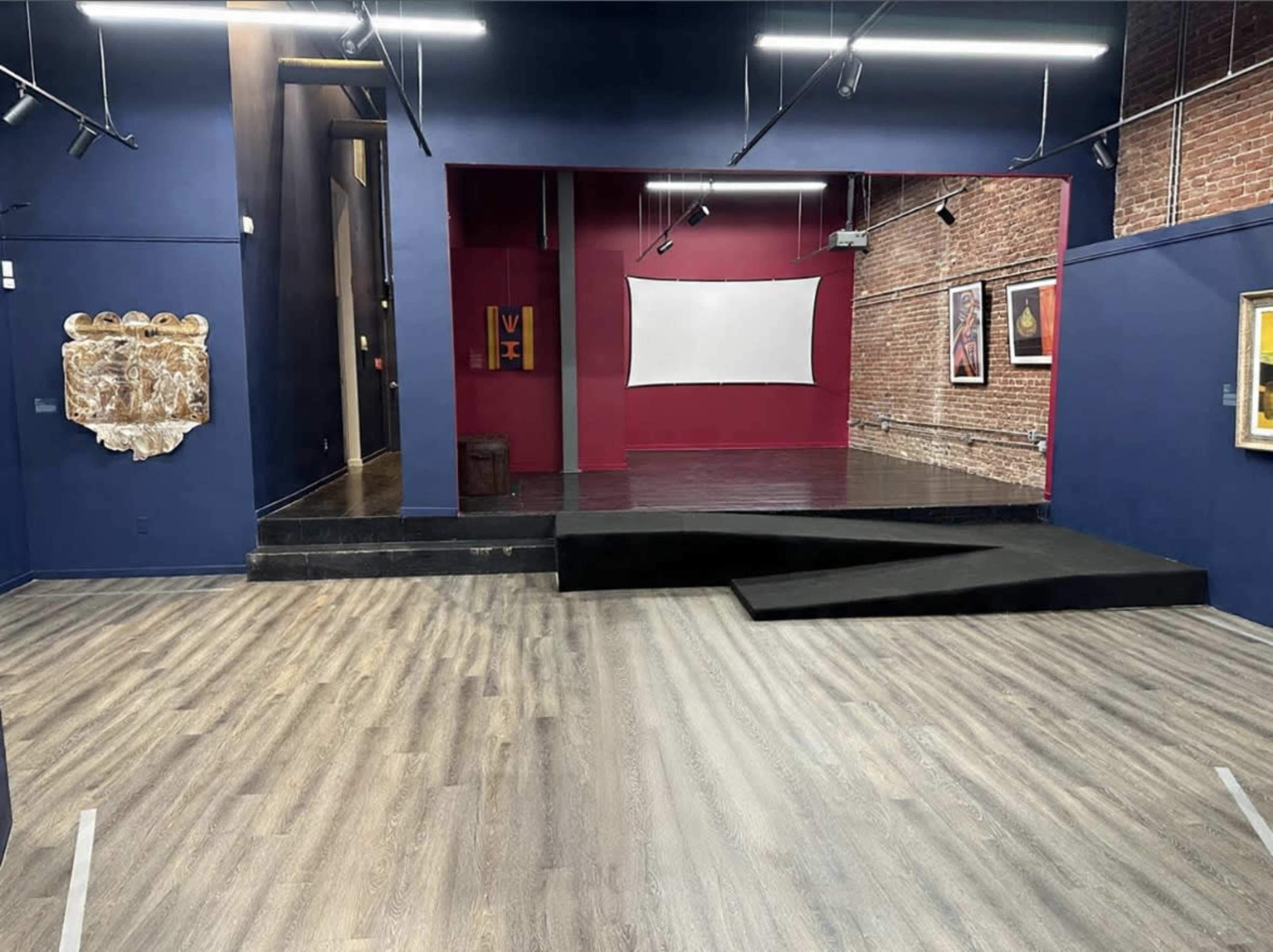 The image shows an interior space with a stage, a large white screen, exposed brick walls, and art displayed on the walls.