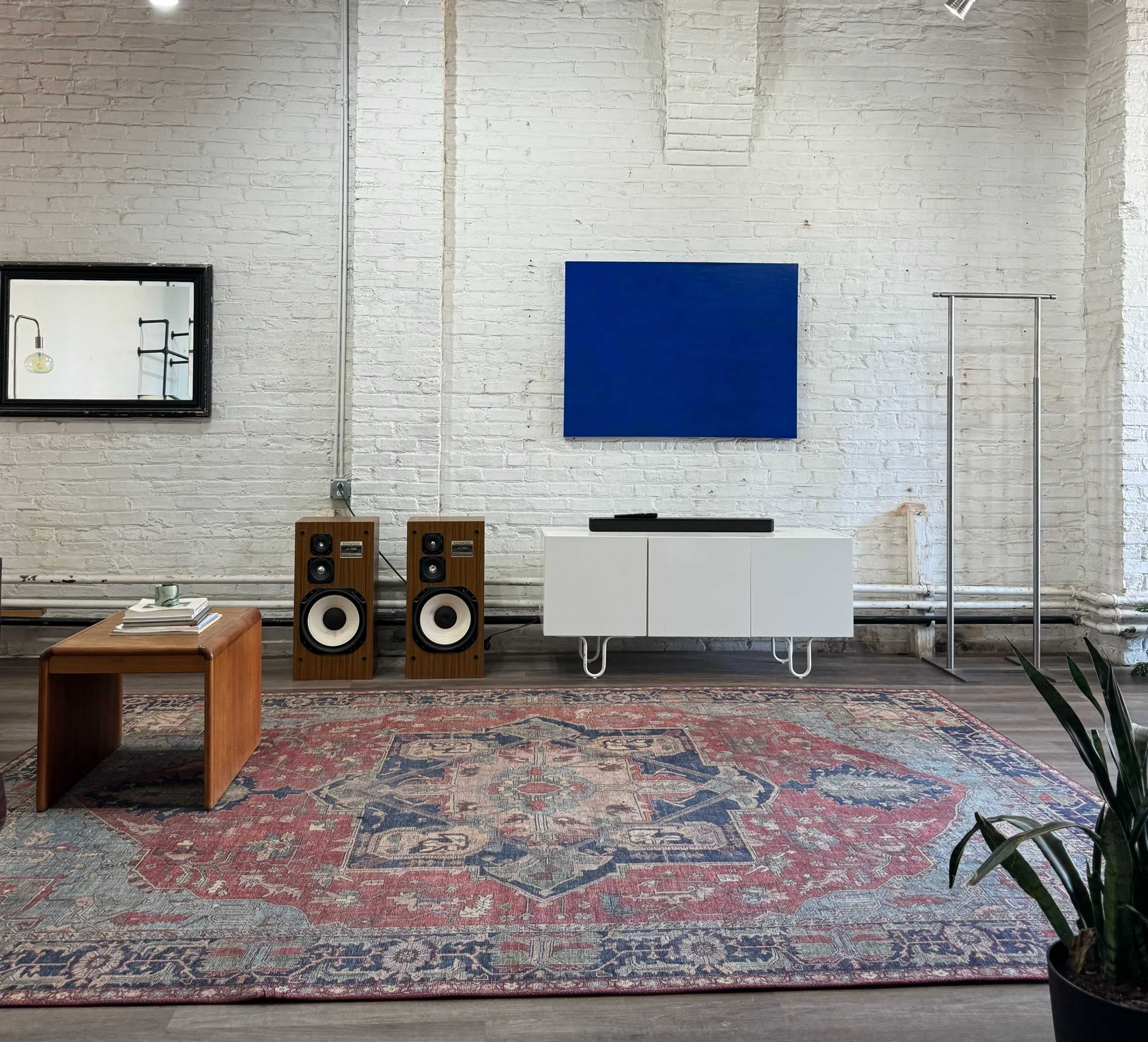 The interior features a white brick wall, a wooden coffee table, a large blue canvas on the wall, a modern media console, vintage speakers, and a patterned area rug.