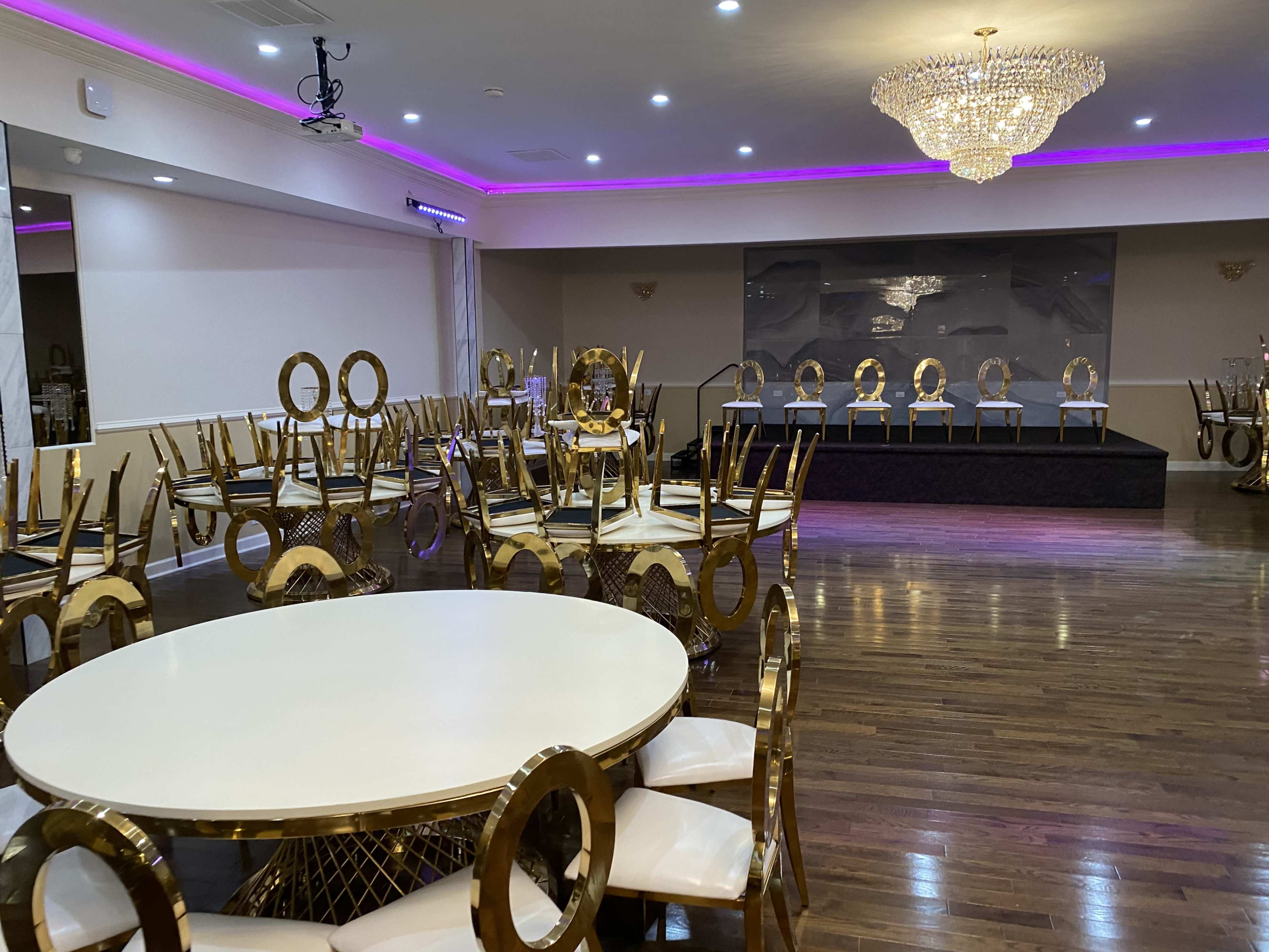 A banquet hall features round tables with gold accents and a raised stage at the far end, illuminated by a large chandelier and purple lighting.