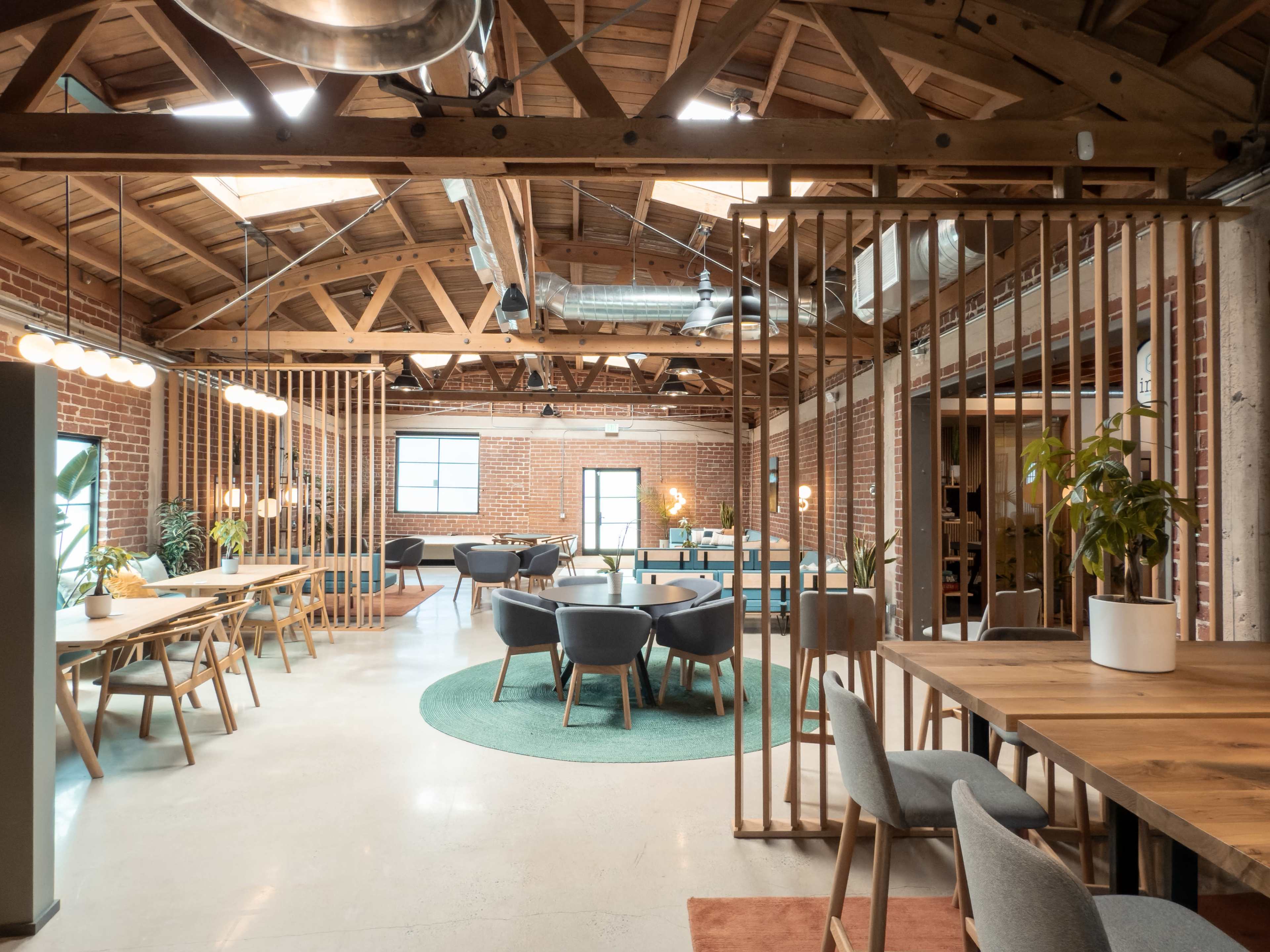The image shows a spacious, modern office interior with wooden beams, a variety of seating areas, and greenery.