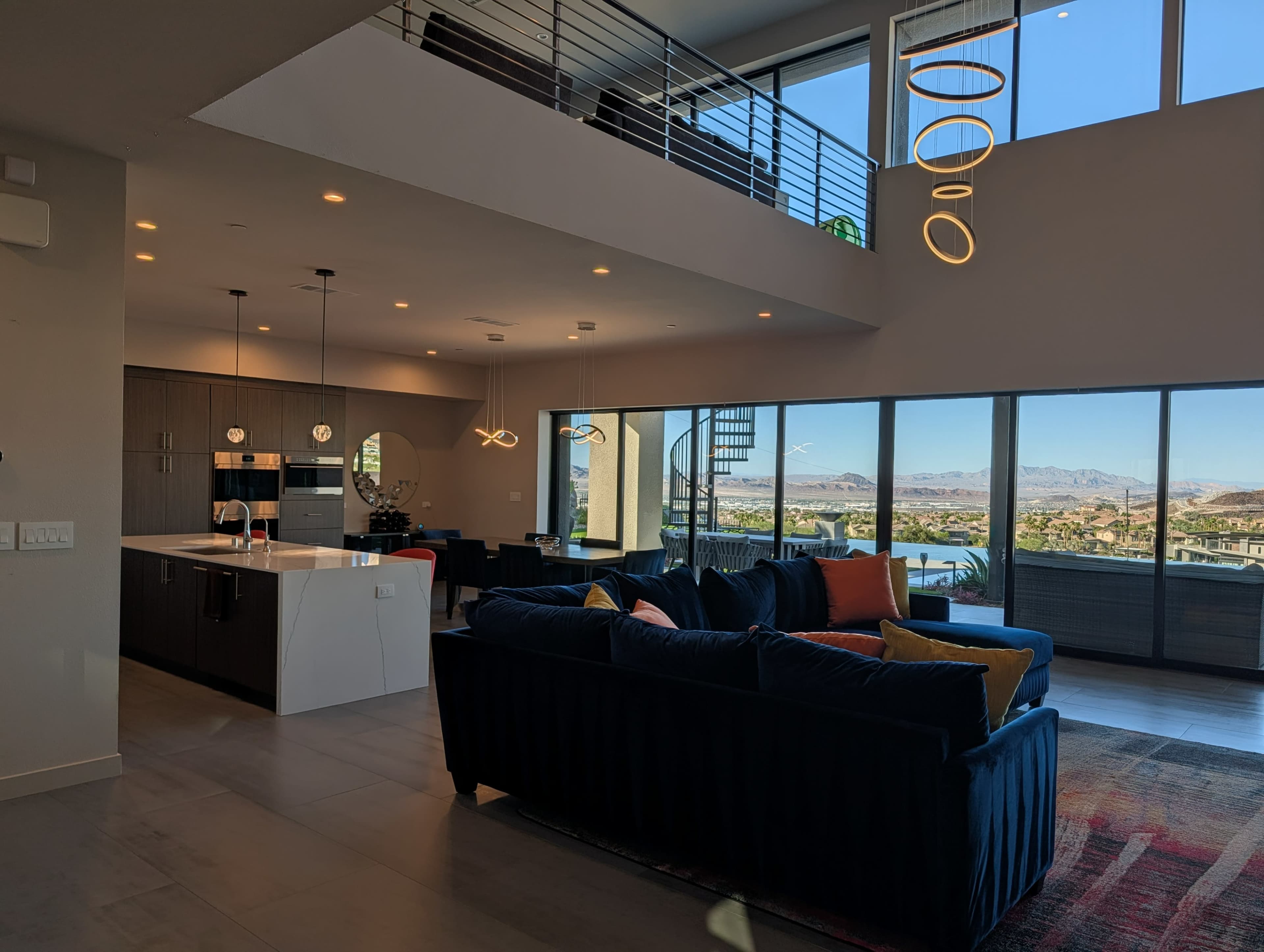 The image shows a modern open-concept living space featuring a stylish kitchen, a plush sofa with colorful cushions, and large windows offering a view of the landscape outside.