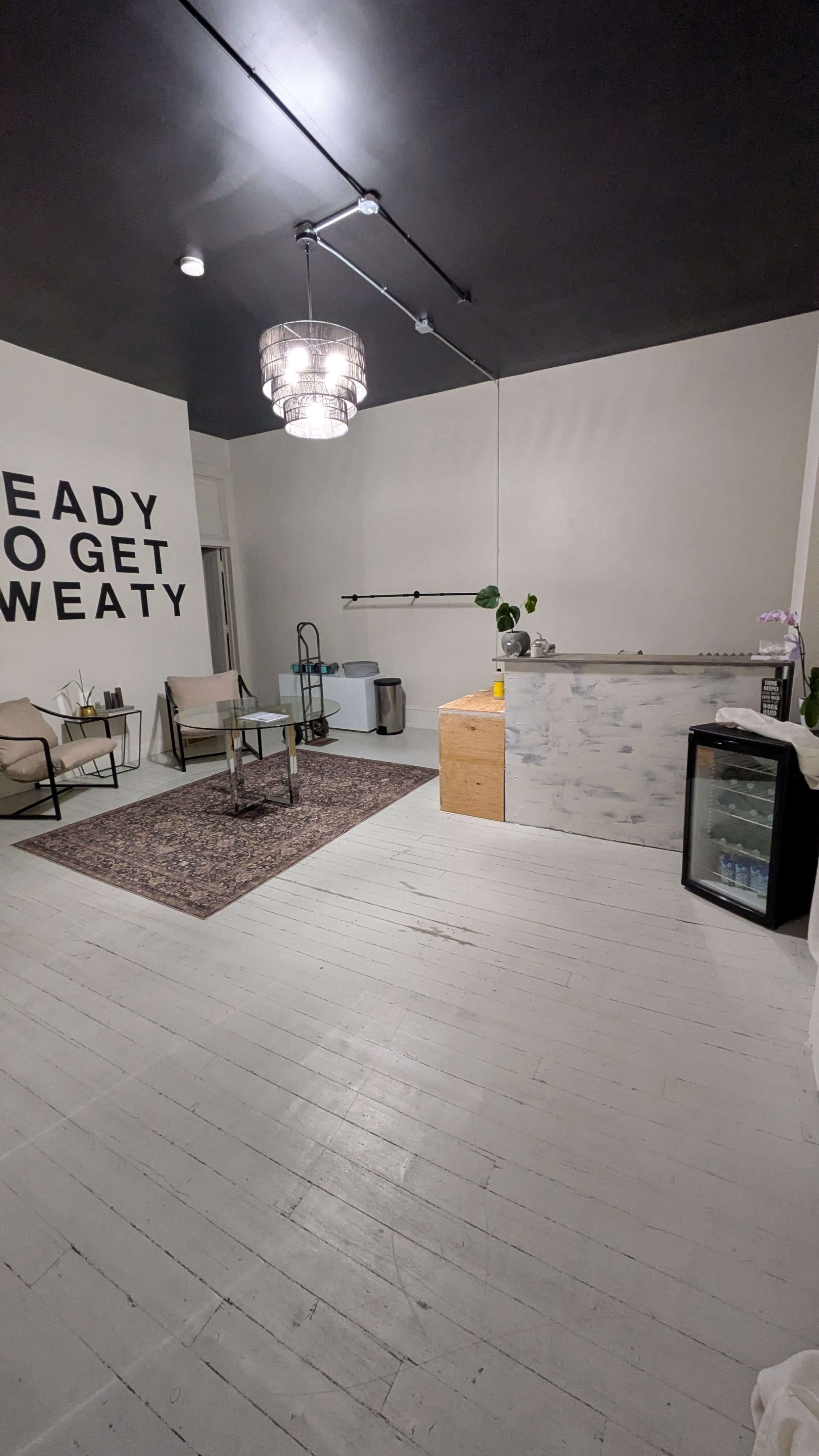 The image shows a spacious room with light wooden flooring, two chairs and a small table, a decorative rug, a reception desk, and a wall featuring the text "READY TO GET SWEATY."