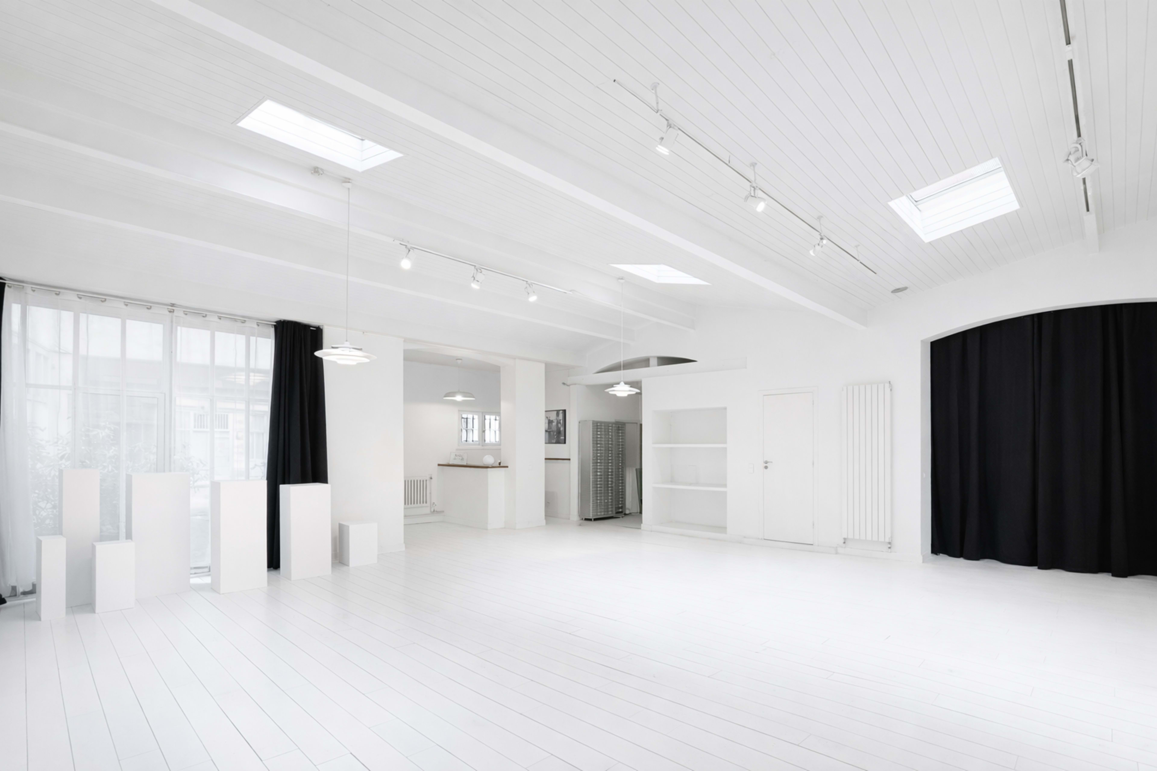 The image shows a spacious, well-lit studio with white walls and wooden floors, featuring large windows and minimal decor.