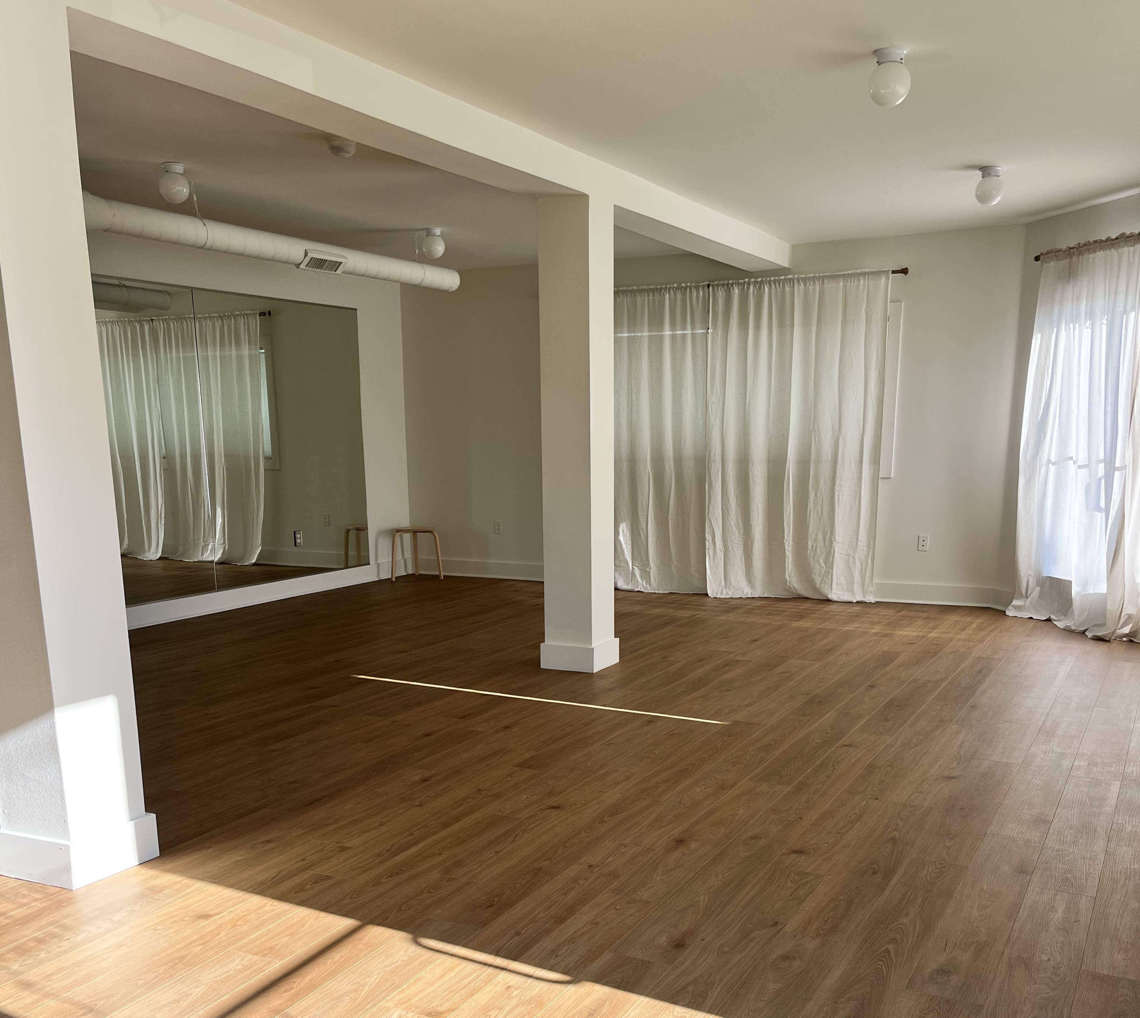 A spacious, empty room features wooden flooring, large windows with sheer curtains, and a full-length mirror on one wall.