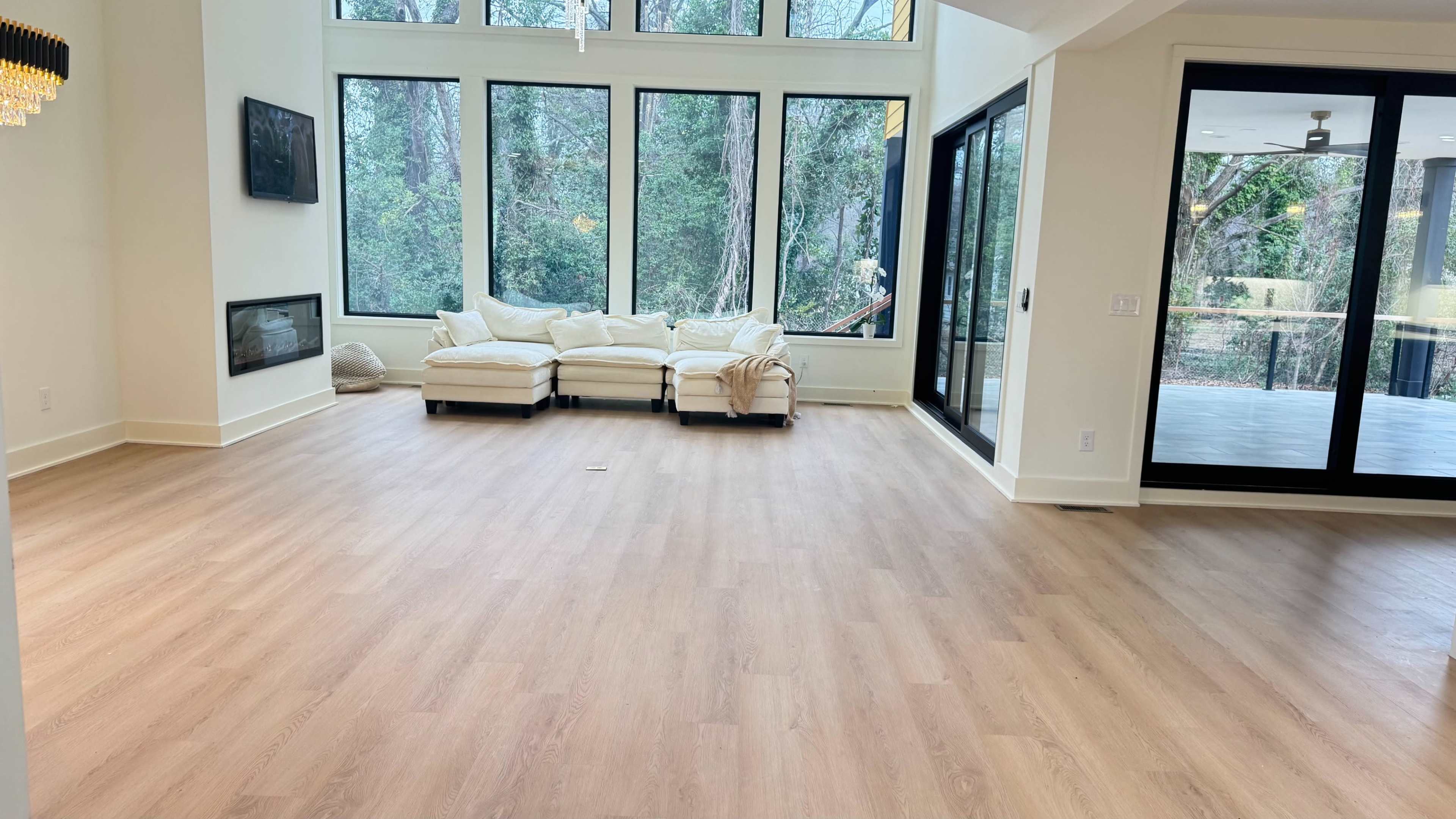 The image shows a spacious, bright room with large windows overlooking a wooded area, featuring a light-colored sofa positioned against the wall.