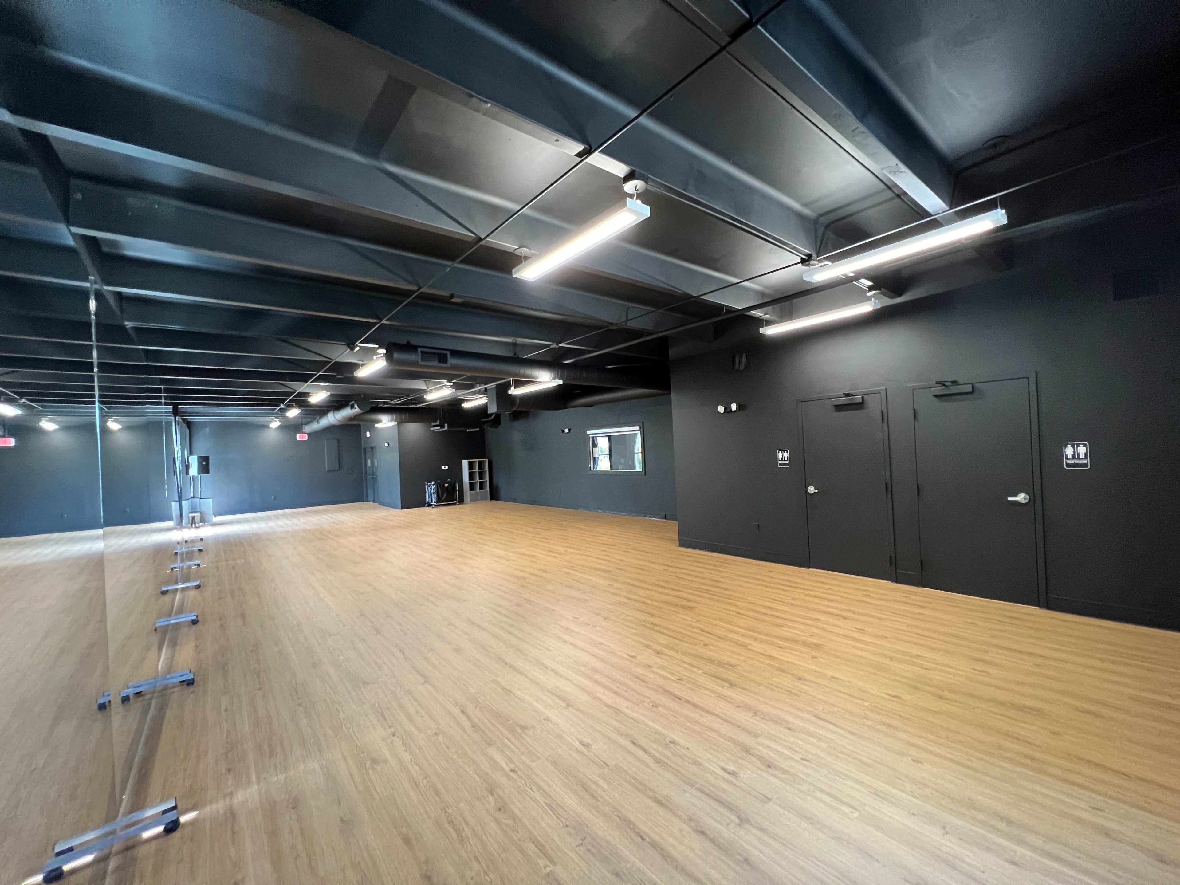 A spacious, empty dance studio featuring a wooden floor, black walls, and a large mirror along one side.