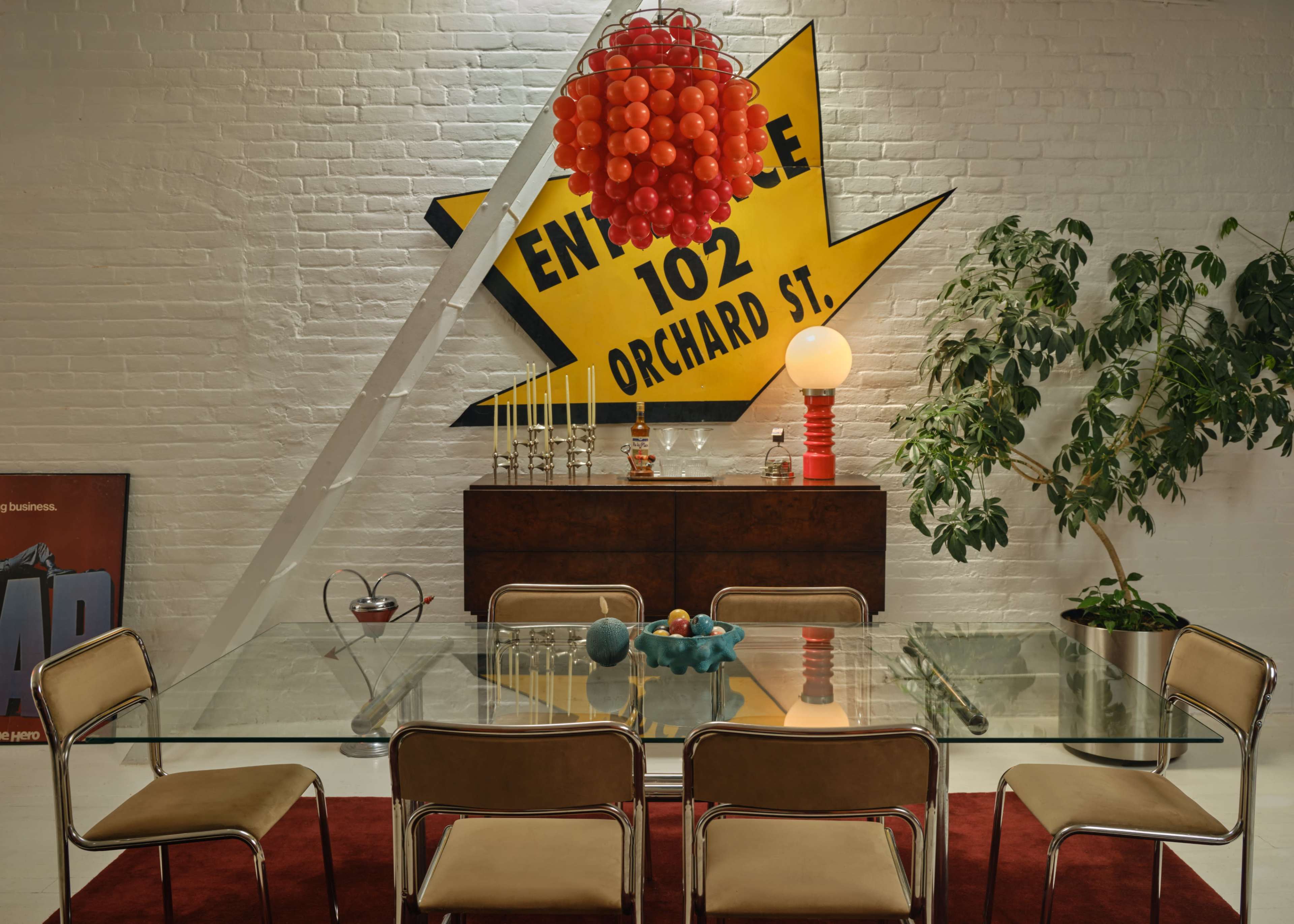 The image shows a modern dining area featuring a glass table surrounded by beige chairs, a colorful pendant light, and a large bold wall sign.