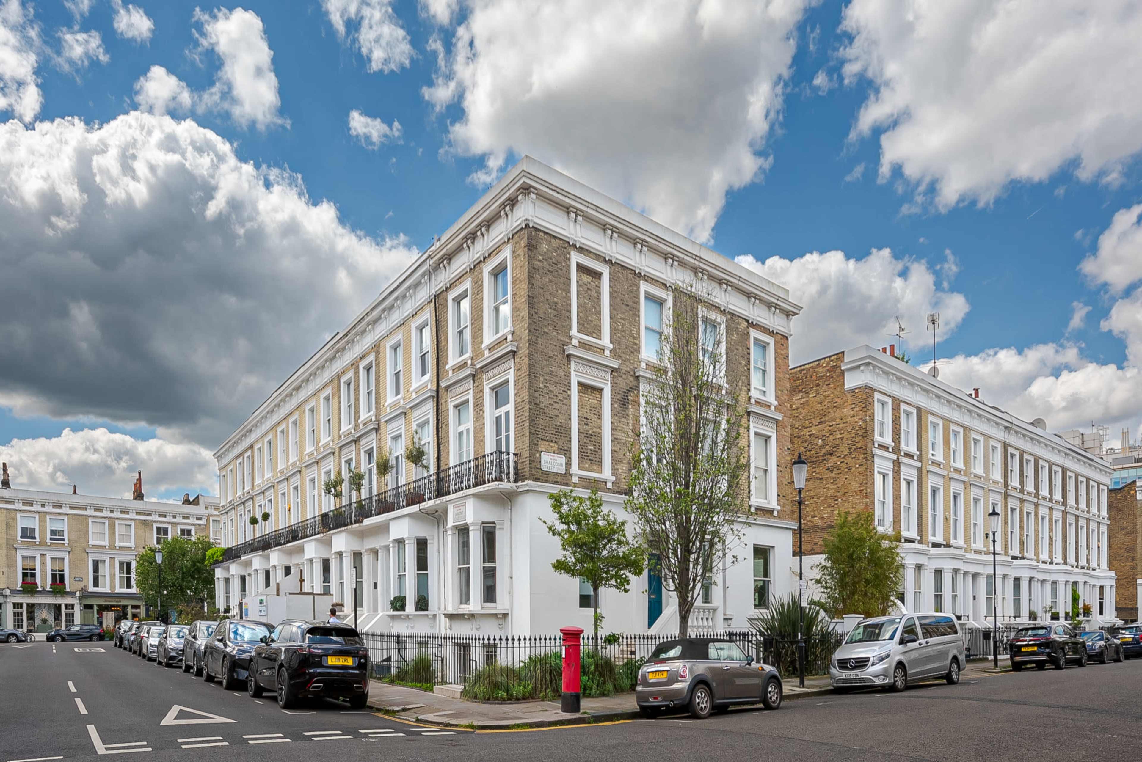 The image shows a street scene featuring several multi-story residential buildings with white façades and balconies, parked cars along the curb, and a red post box in the foreground under a cloudy sky.