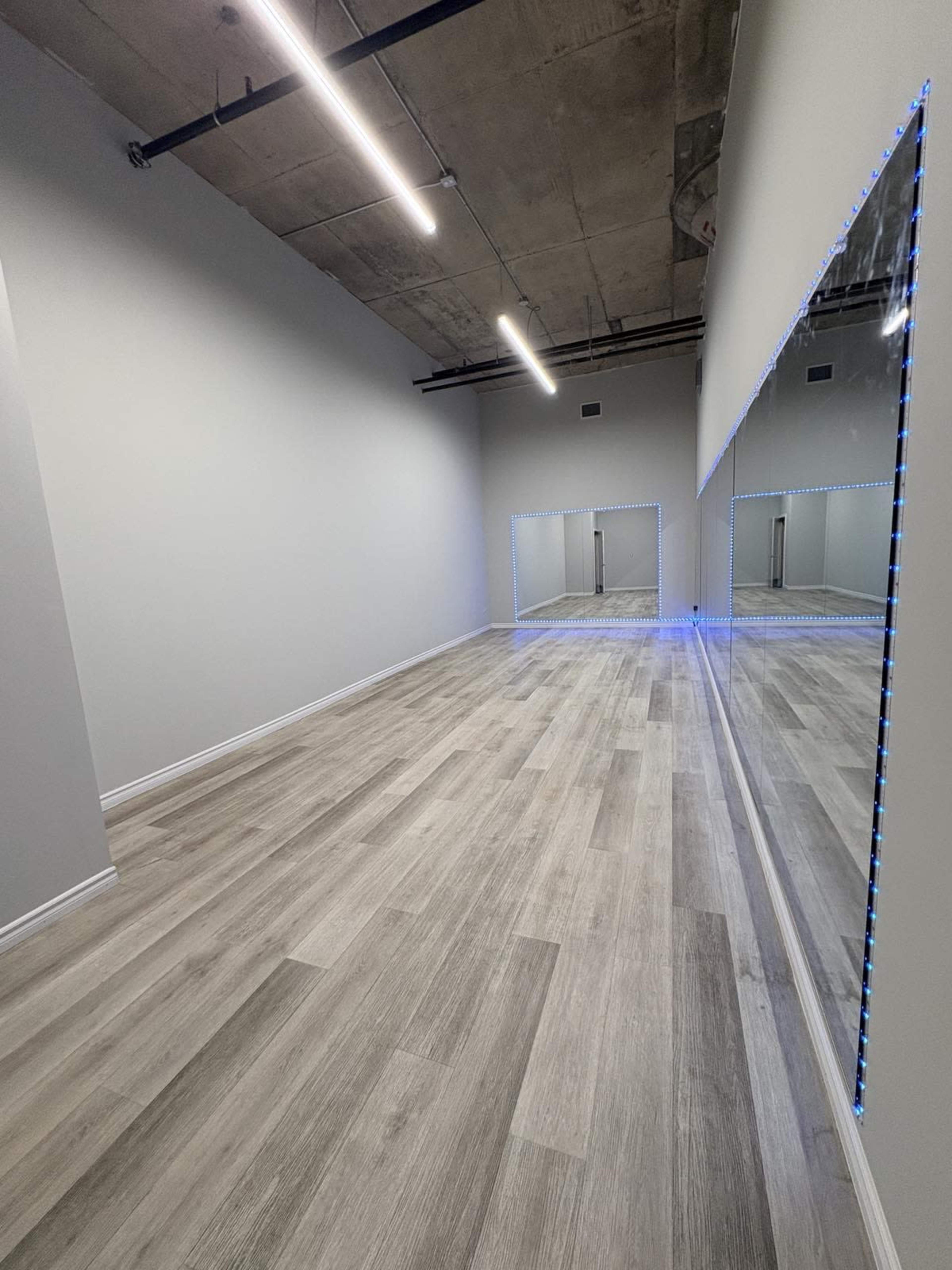 The image shows a spacious, empty room with light-colored wooden flooring and mirrored walls, illuminated by fluorescent lights.