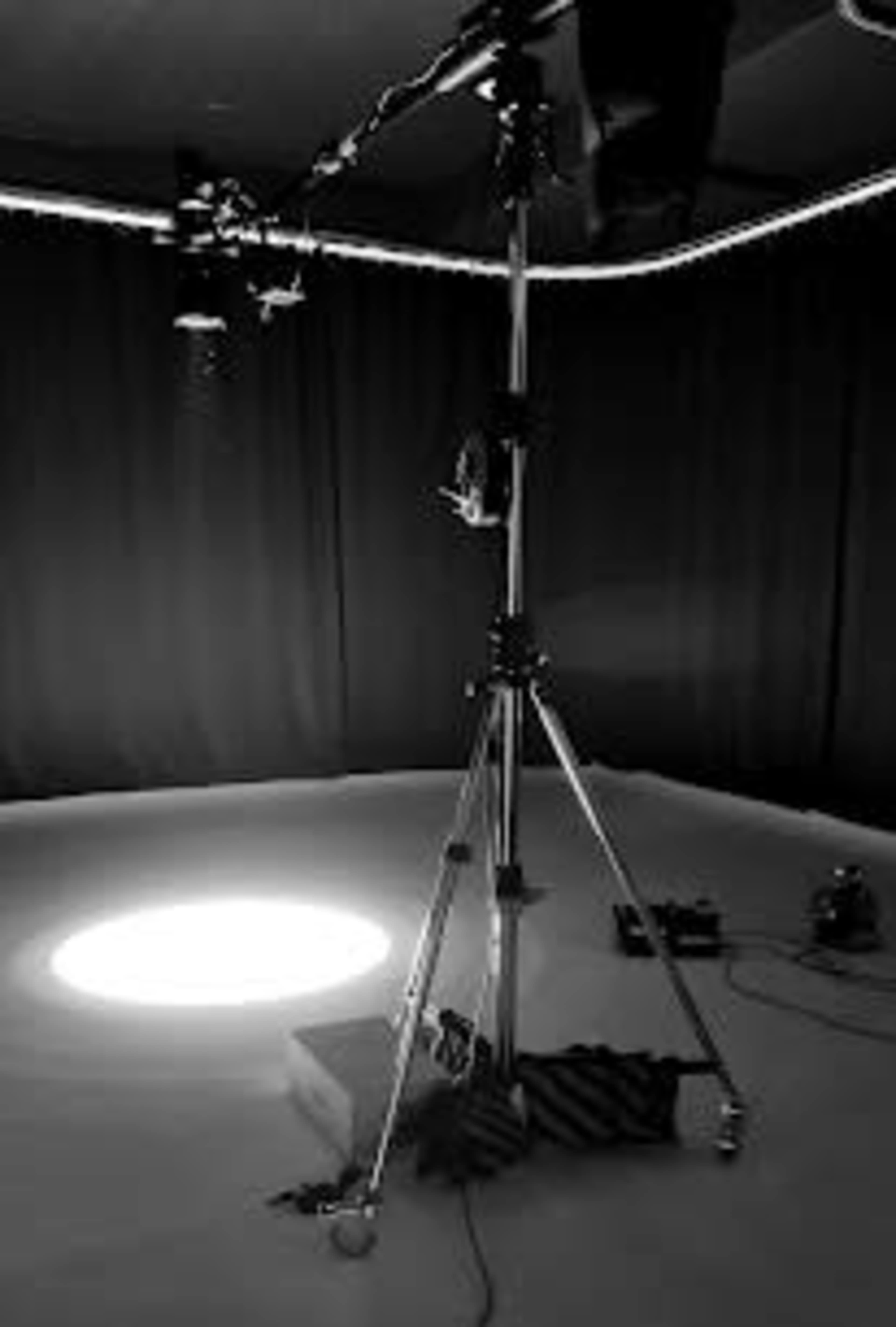 A camera tripod stands in a dimly lit room, casting a spotlight on the floor.