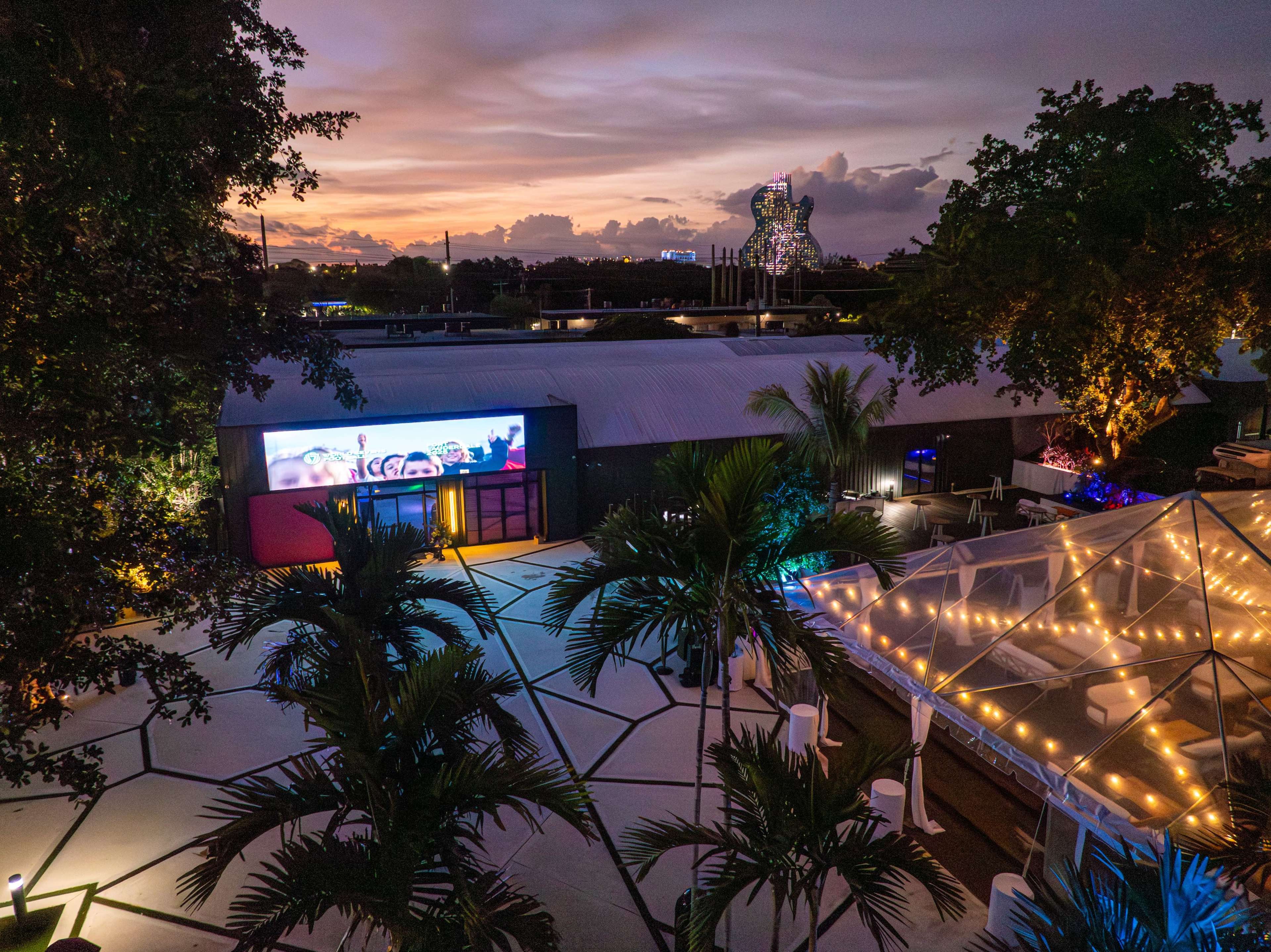 The image shows a landscaped outdoor area with a tent illuminated by string lights, a large screen displaying colorful visuals, and a sunset in the background.