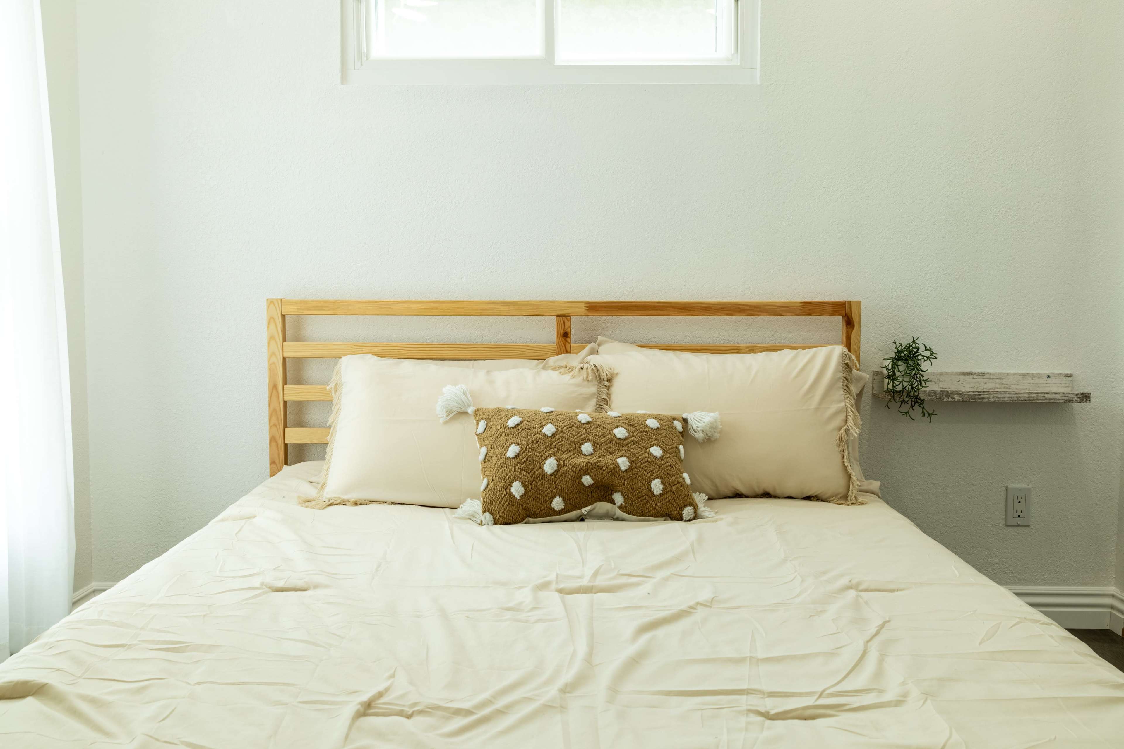 A neatly made bed with a wooden headboard, beige bedding, and decorative pillows, set against a light-colored wall with a window.
