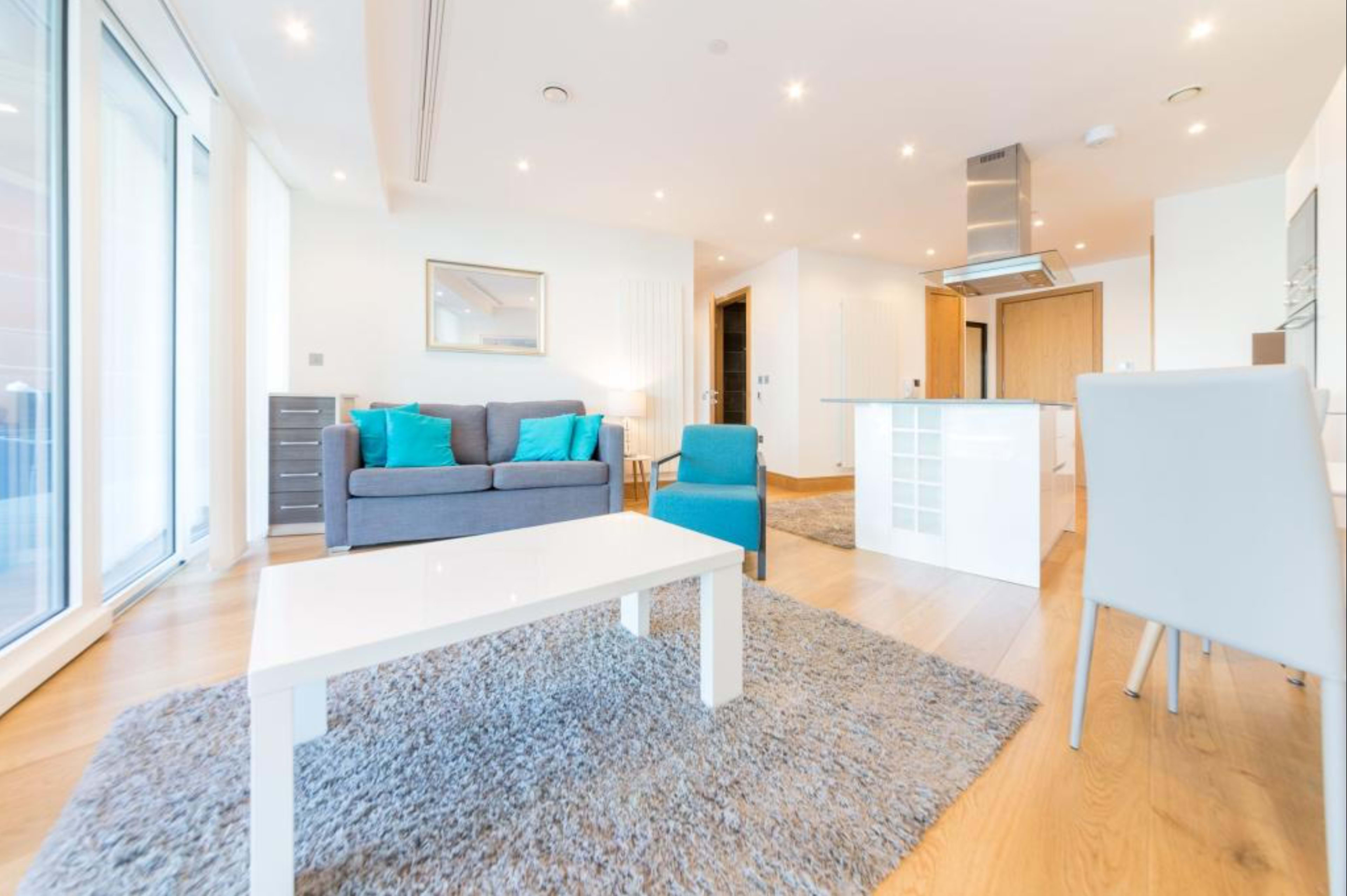 A modern living room features a gray sofa and a light blue chair, with a white coffee table and a rug on a wooden floor, leading to a bright kitchen area.