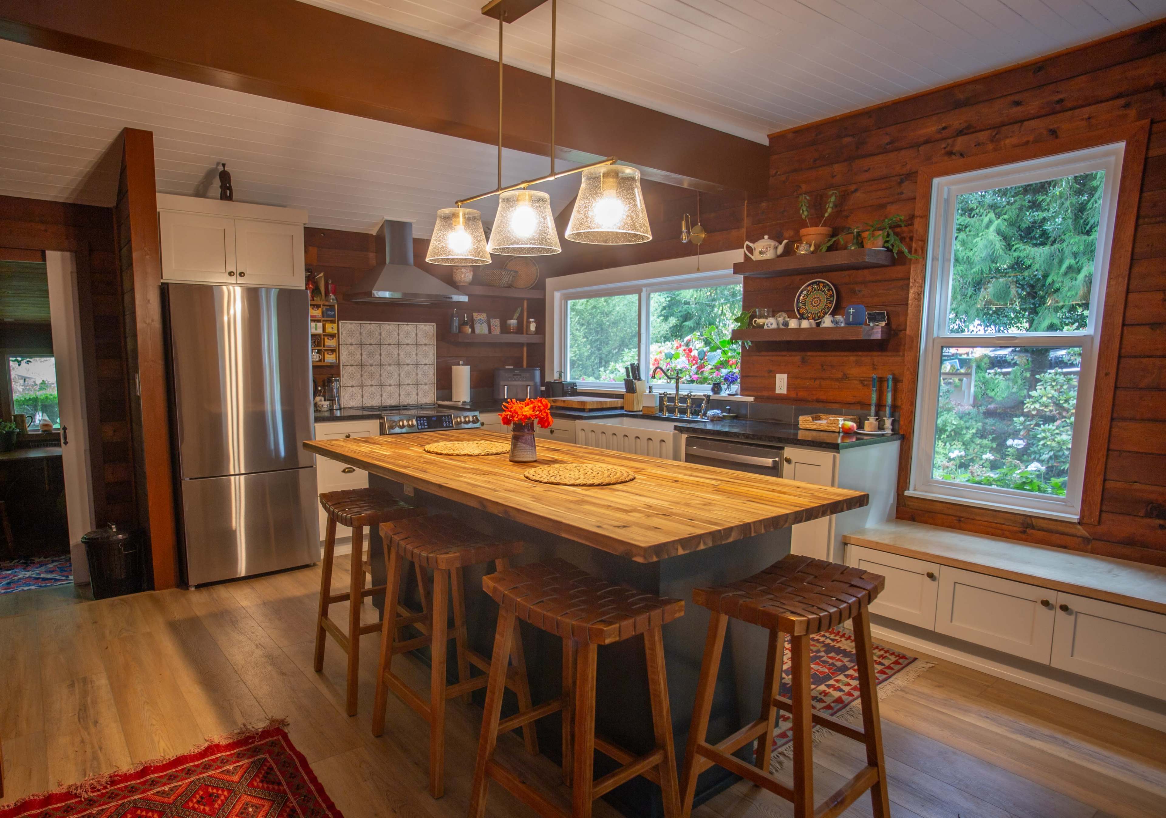 The kitchen features wooden walls, a central island with bar stools, and large windows that overlook a landscaped yard.