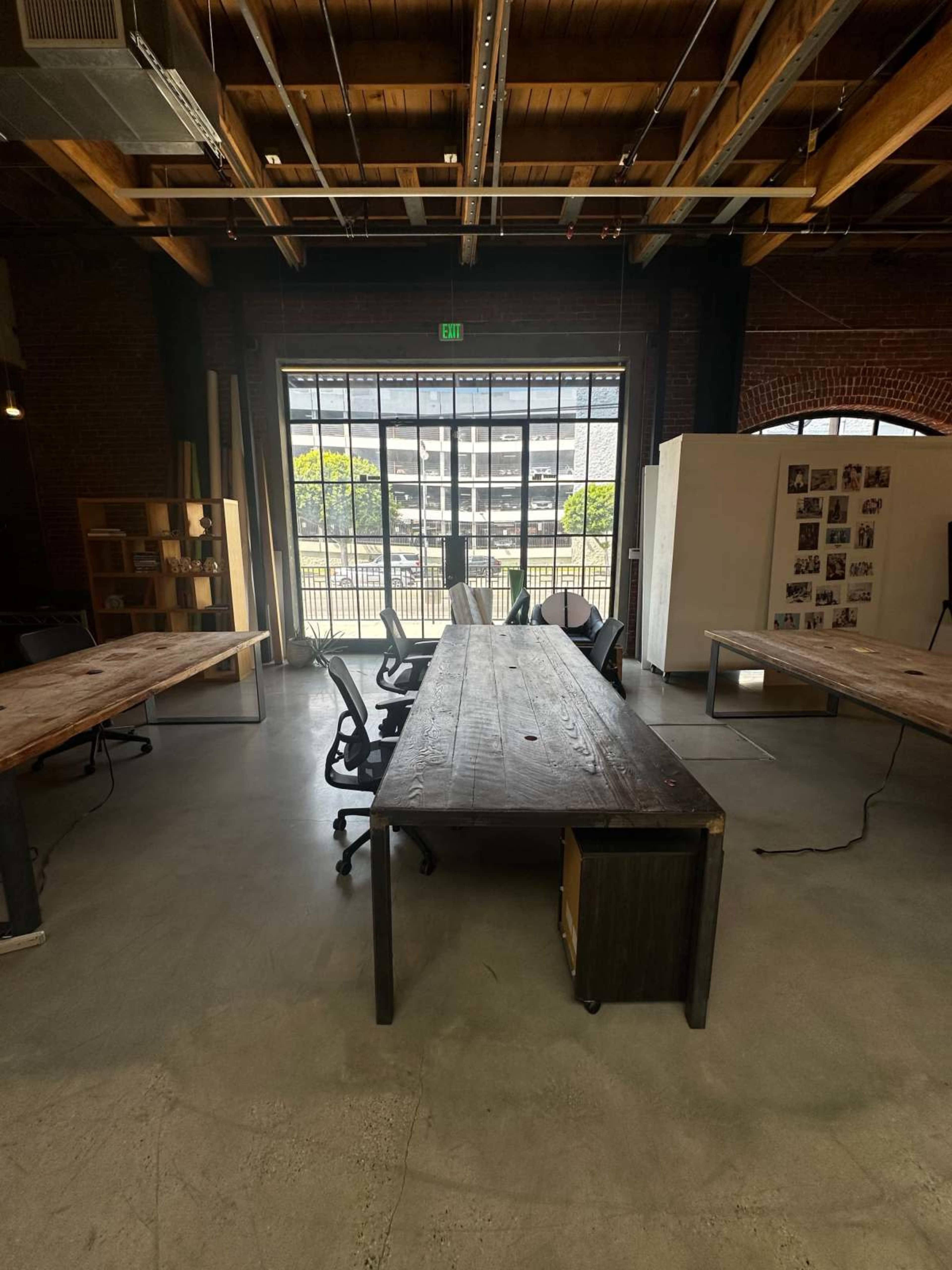 A long wooden table with office chairs is positioned in a bright, spacious room featuring large windows and exposed wooden beams.