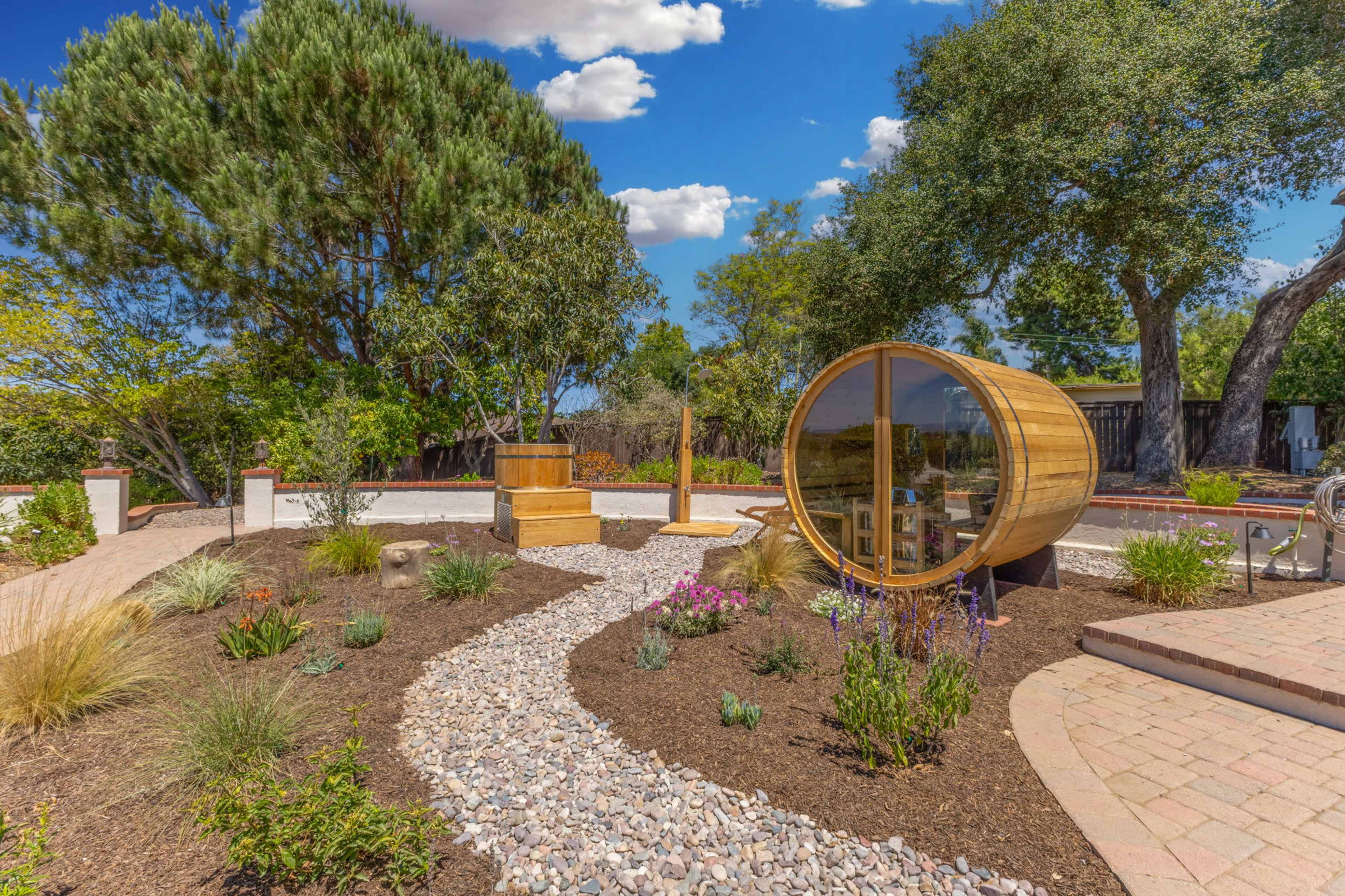 The scene features a landscaped garden with a winding gravel path, a wooden sauna barrel, a wooden hot tub, and various plants under a blue sky with scattered clouds.