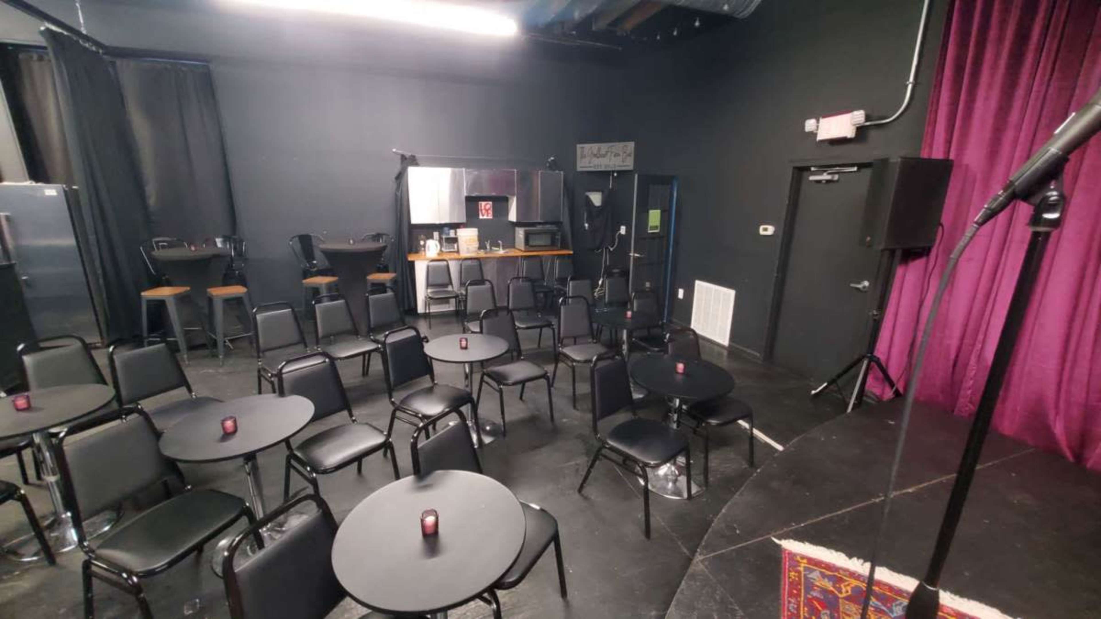 The image shows a small performance space set up with several black tables and chairs arranged in front of a stage, with a purple curtain in the background.