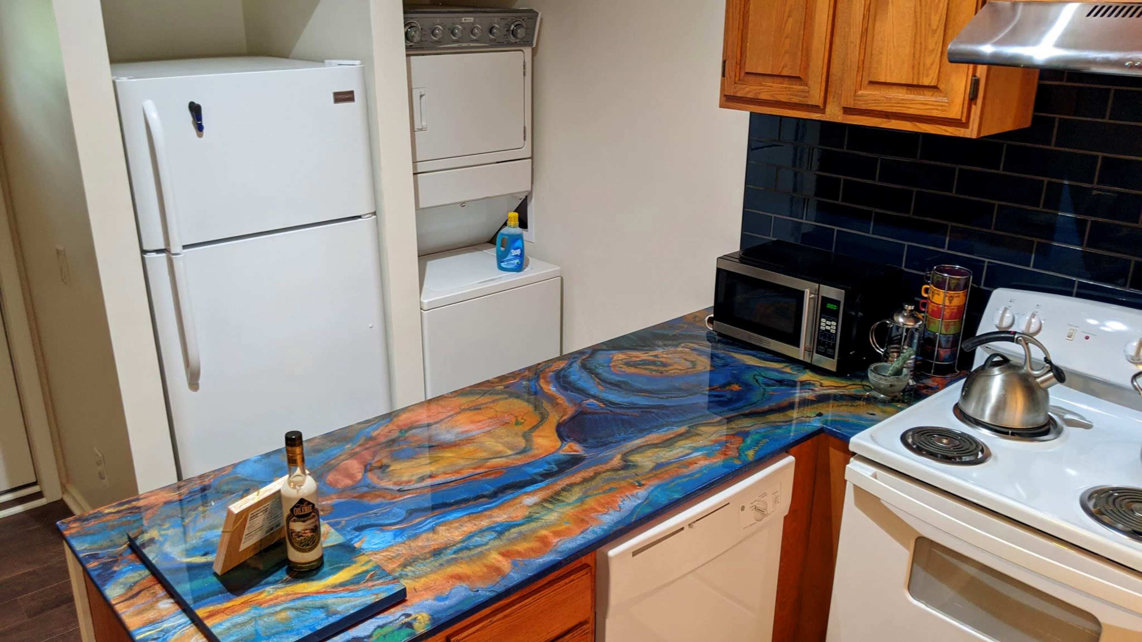 The image shows a modern kitchen with wooden cabinets, a blue and orange patterned countertop, a white refrigerator, a washing machine in the adjacent area, and various kitchen appliances including a microwave and a stove.
