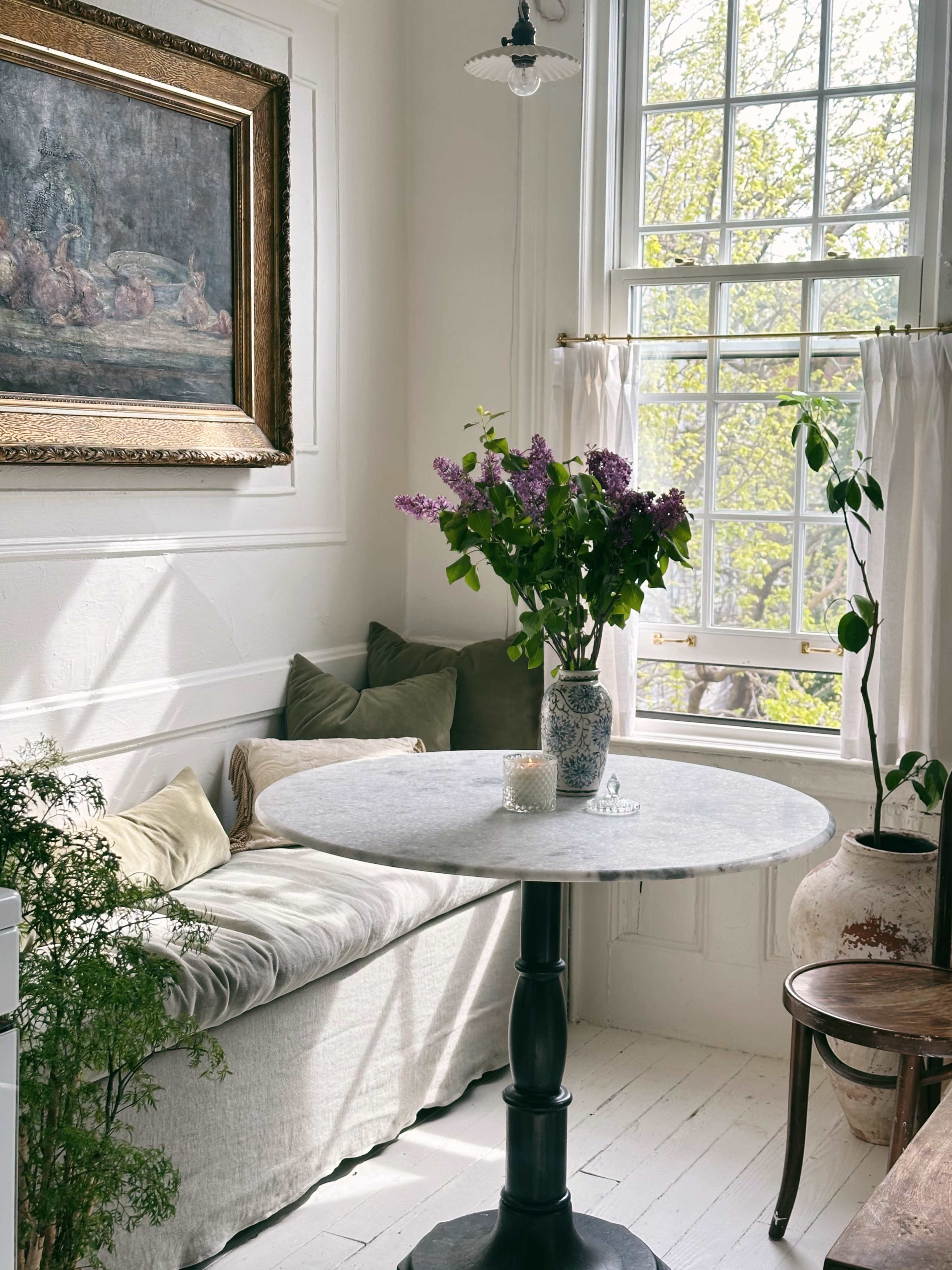 A round marble table with a vase of lilacs is positioned beside a window in a bright, airy room featuring a couch and a framed painting on the wall.