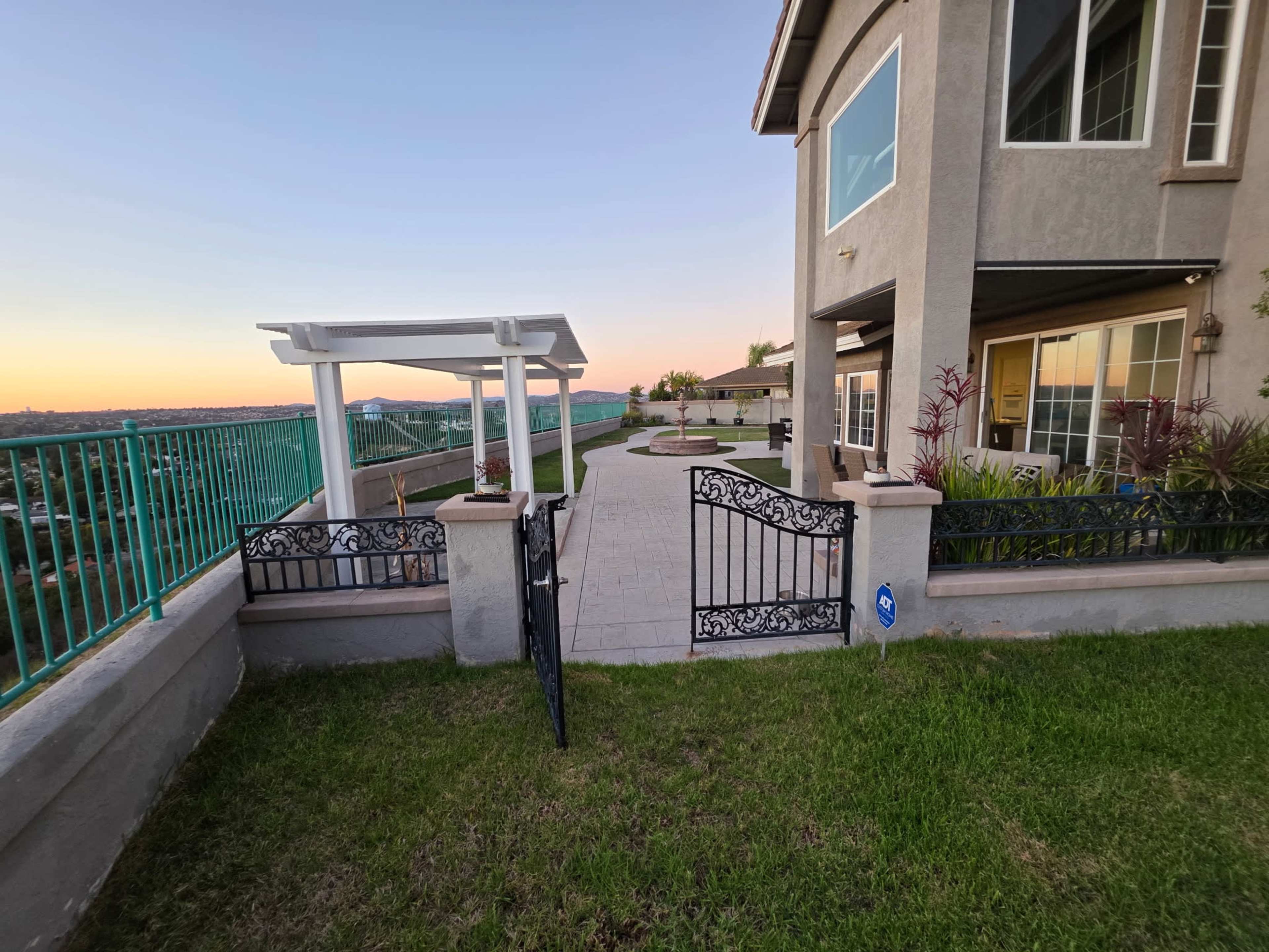 The image shows a landscaped backyard of a house with a gated entrance, a pergola, and an expansive view of the horizon at sunset.