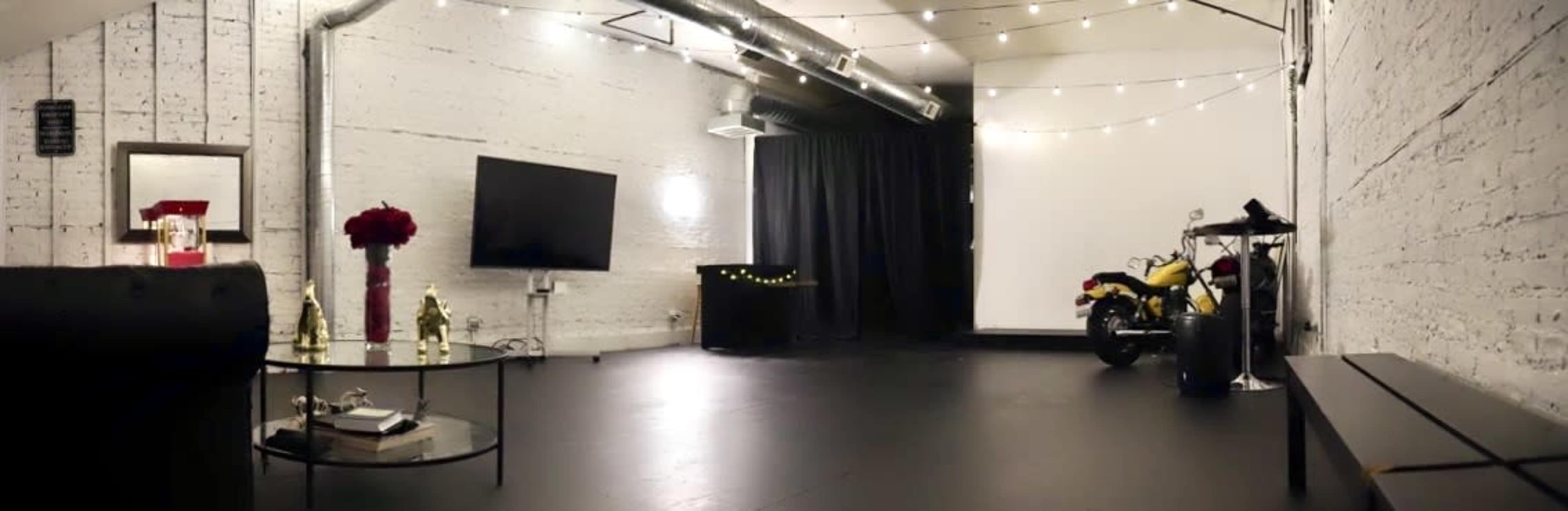 A spacious studio with a black wooden floor, a TV mounted on the wall, a motorcycle parked in the corner, and string lights overhead.