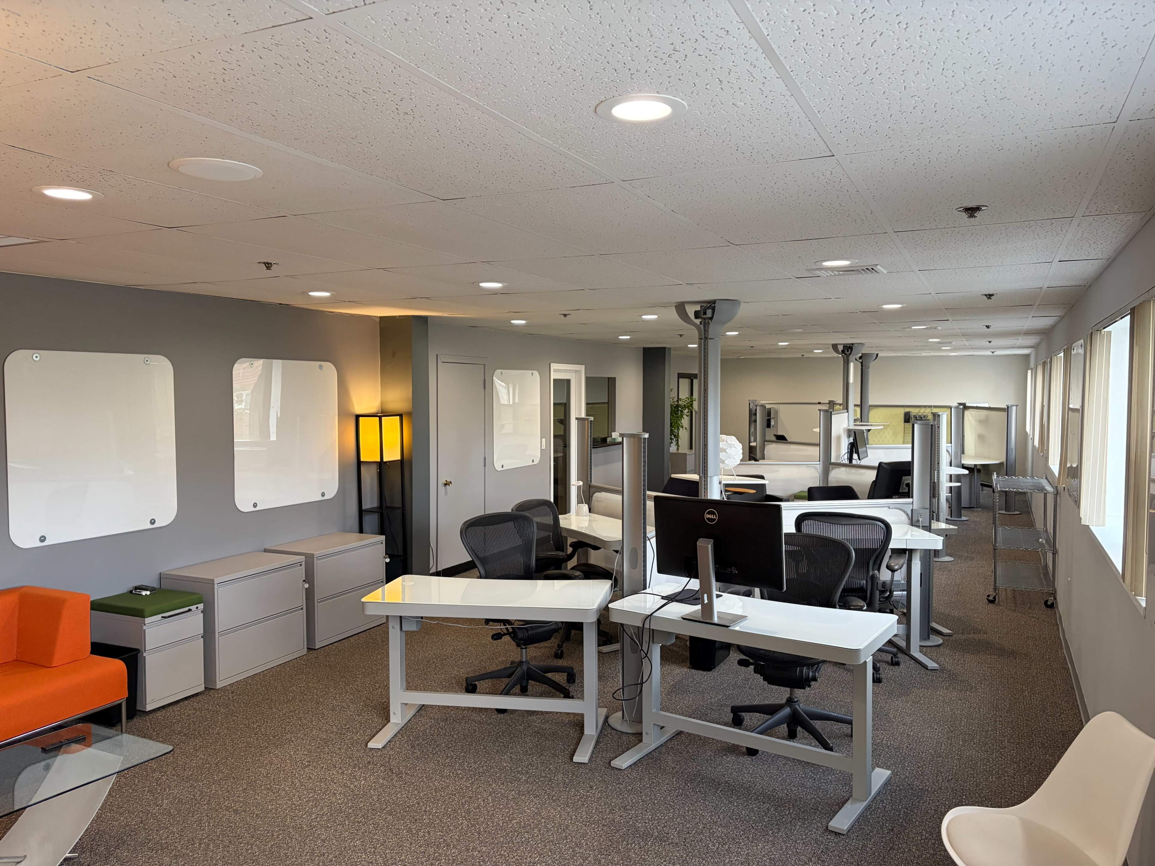 The image shows a modern office space featuring multiple workstations, a seating area with an orange couch, and large windows allowing natural light to enter.