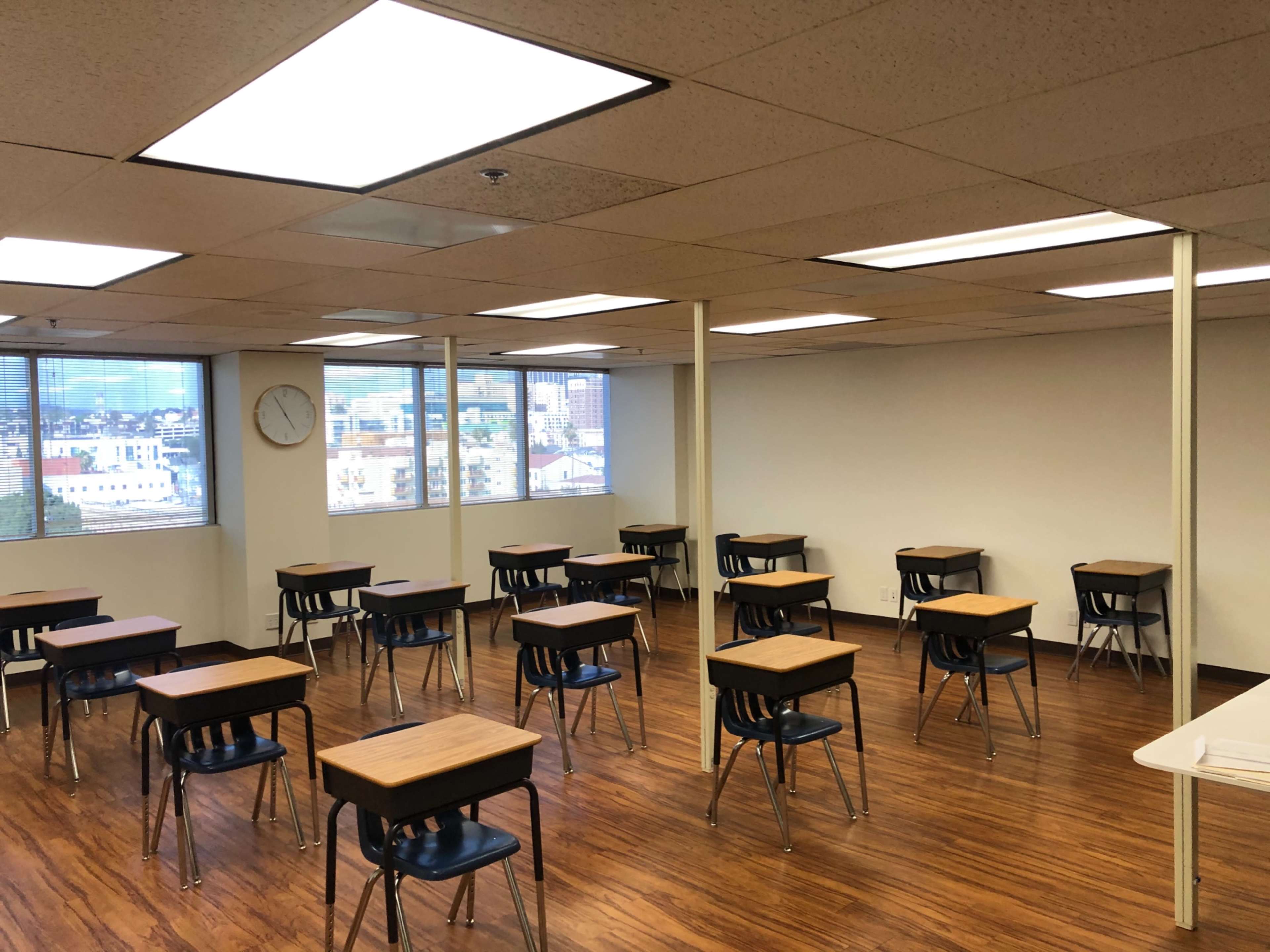 The image shows a classroom with wooden desks arranged in rows, large windows providing natural light, and a clock on the wall.