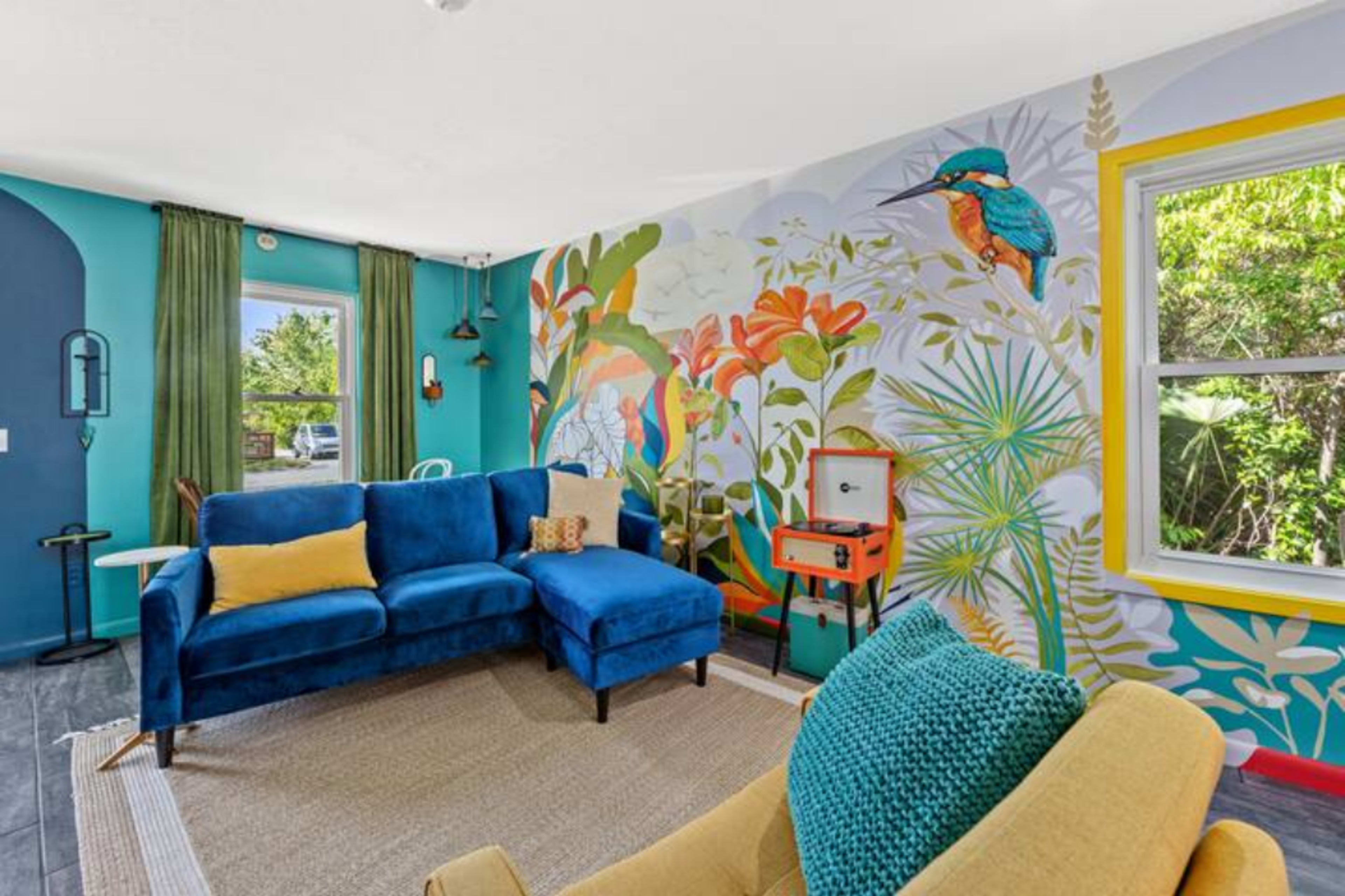 A living room features a blue sectional sofa and a yellow armchair, with a colorful mural of plants and birds on one wall.