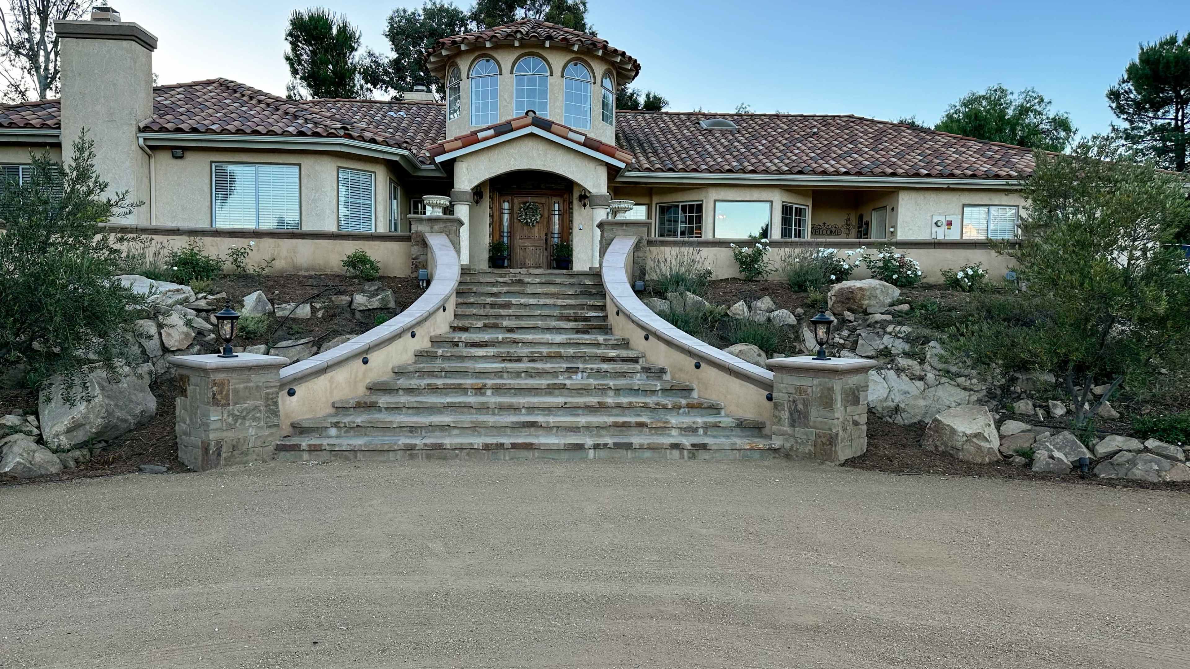 A large house features a front stairway leading up to a rounded entrance, flanked by landscaped greenery and stonework.