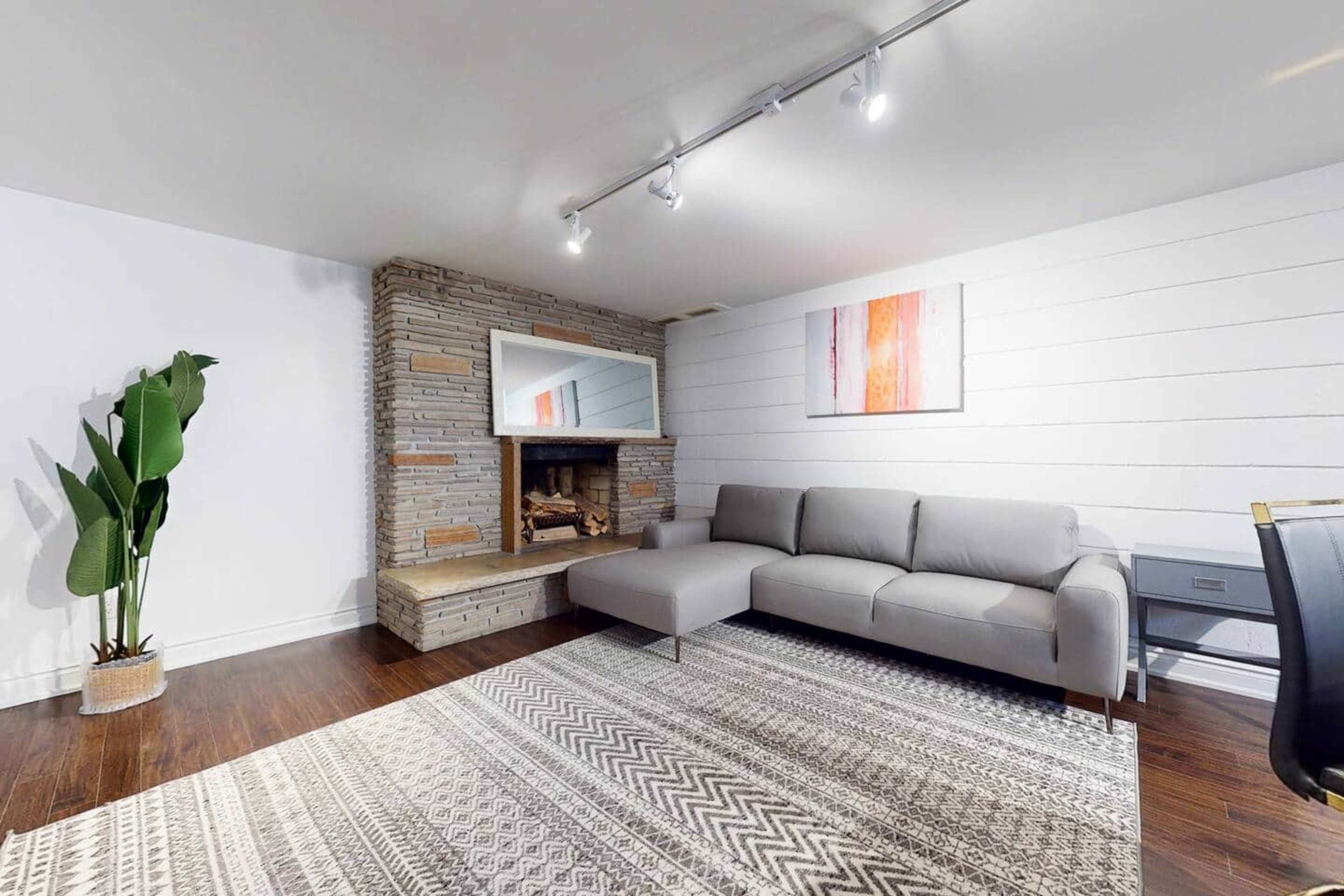 The image shows a modern living room featuring a gray sectional sofa, a stone fireplace, a large area rug, and a leafy plant.