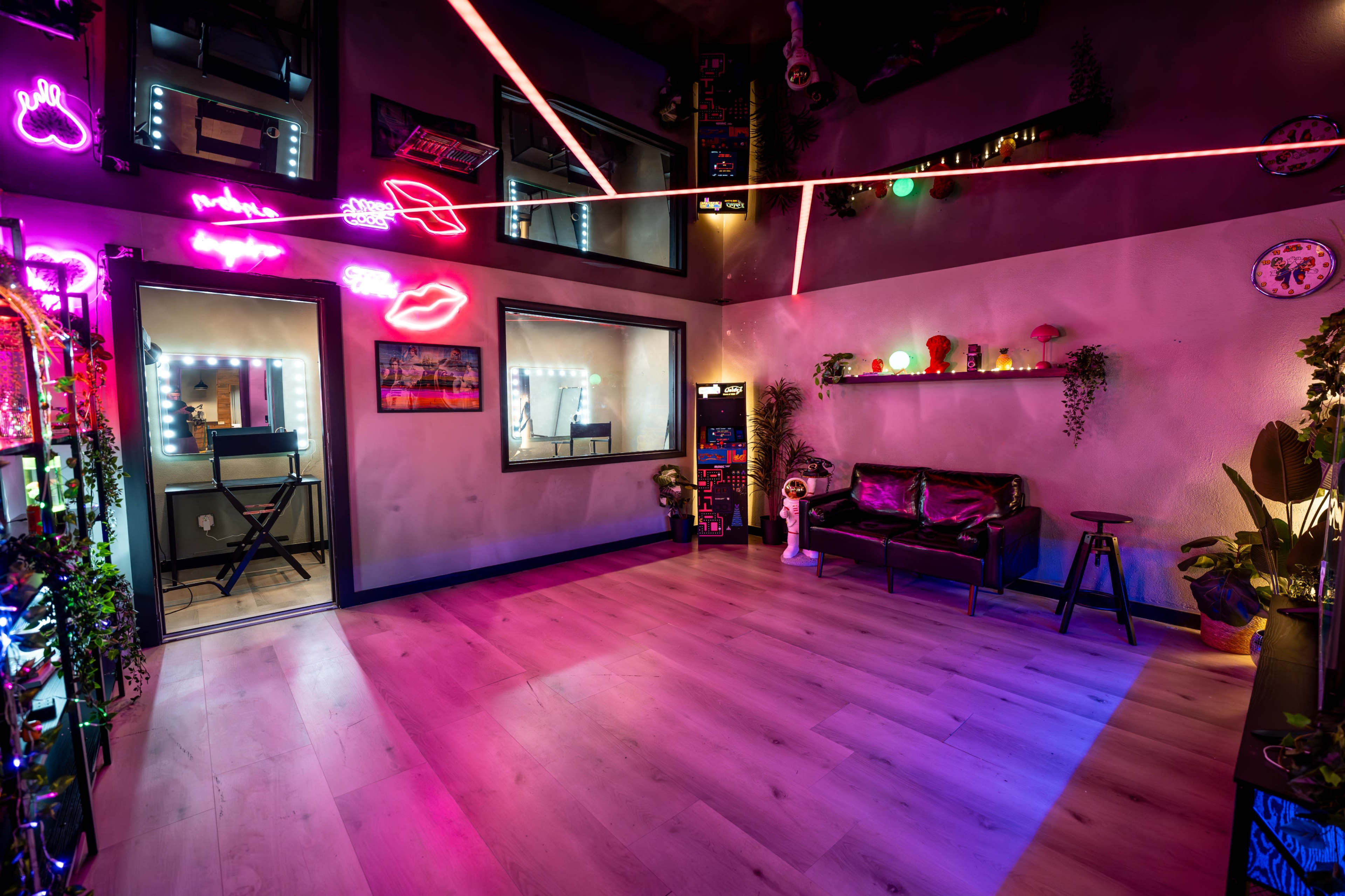 The image shows a brightly lit room featuring neon lights, mirrors, and a modern seating area, with a keyboard in one corner and colorful decor along the walls.